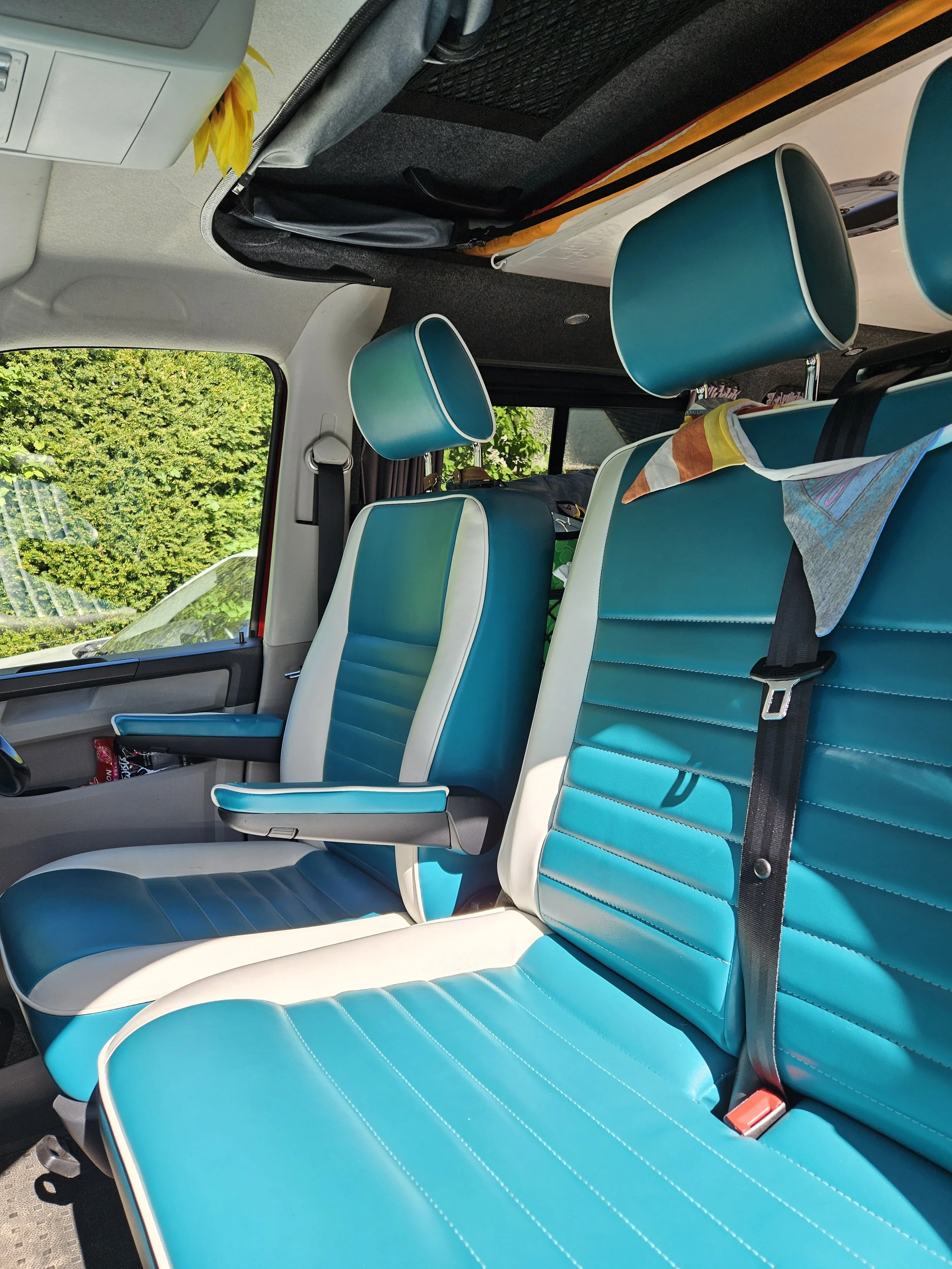 Inside a vehicle with bright blue and white seats, black seatbelts, a window showing greenery outside, and various items stored behind the seats.