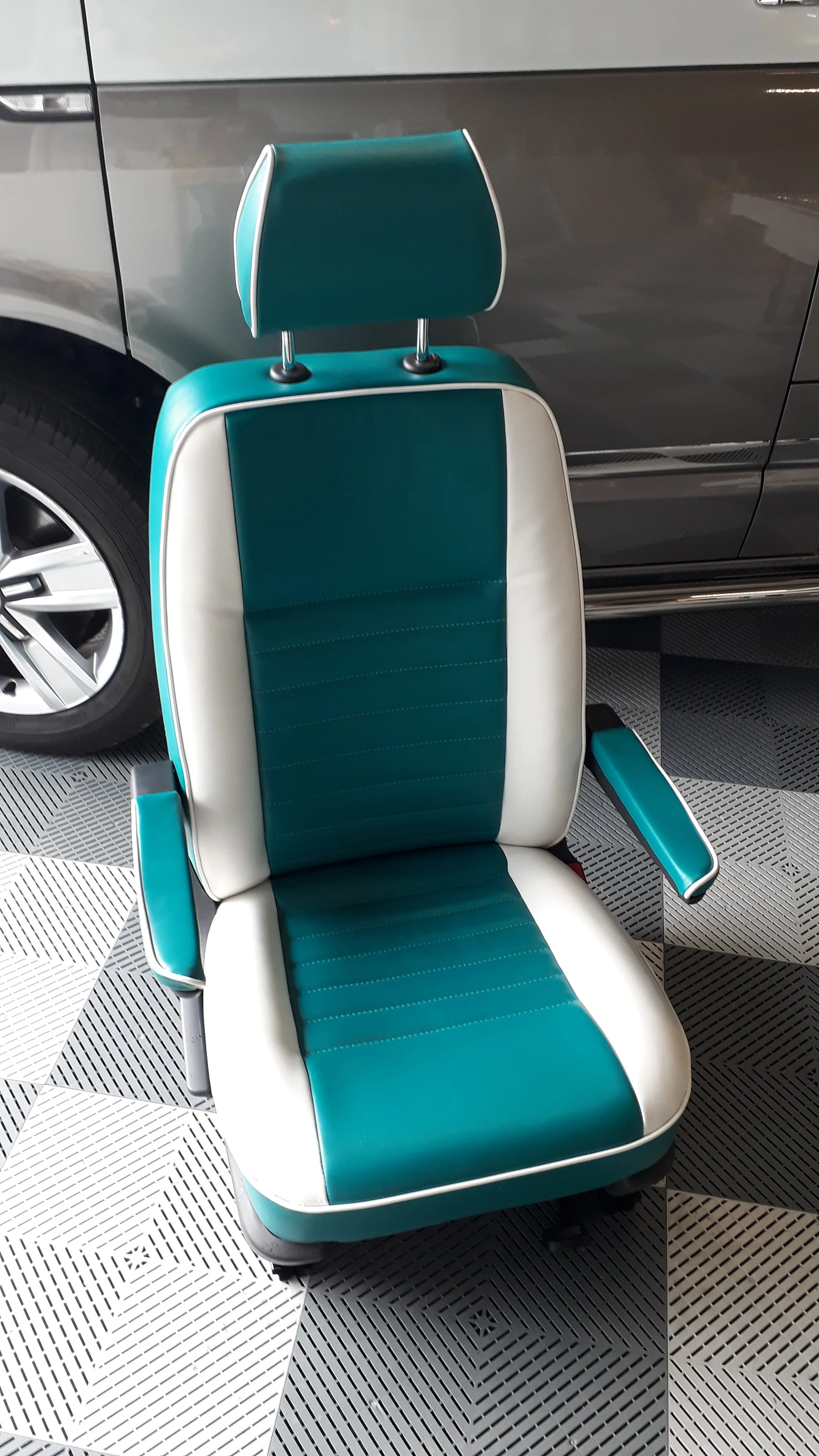 A vintage-style teal and white leather car seat with an adjustable headrest, located in an indoor space with black and white patterned tiled flooring and a gray vehicle in the background.