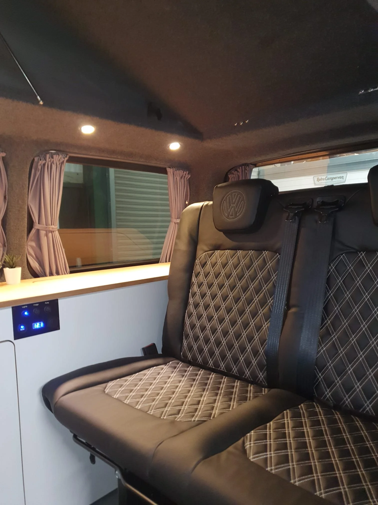 Interior of a camper van or RV with captain's seat, window with pink curtains, and a digital control panel on the wall.