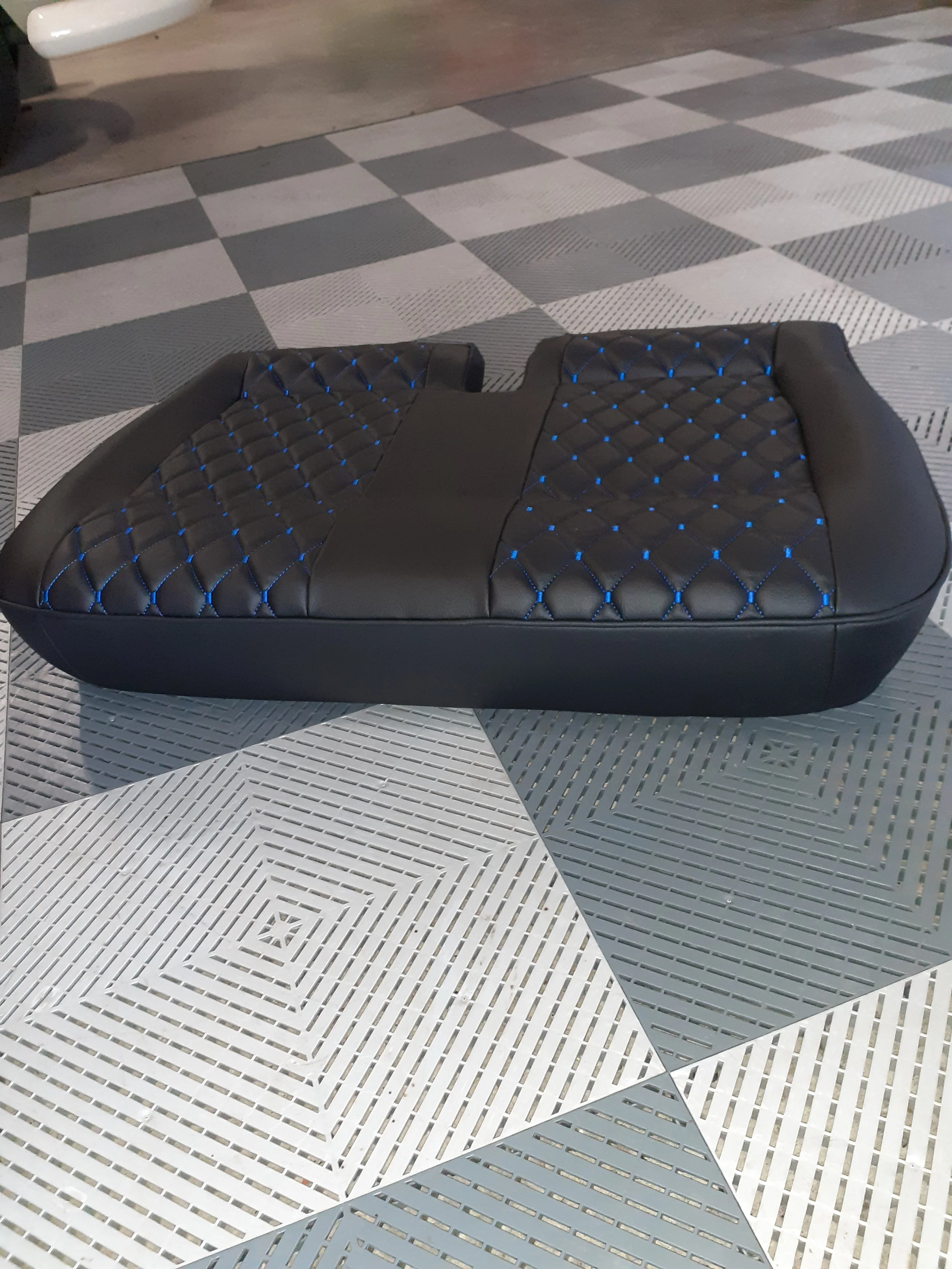 A black, quilted, padded lounge chair with blue stitching on a tiled floor with diamond and square patterns.