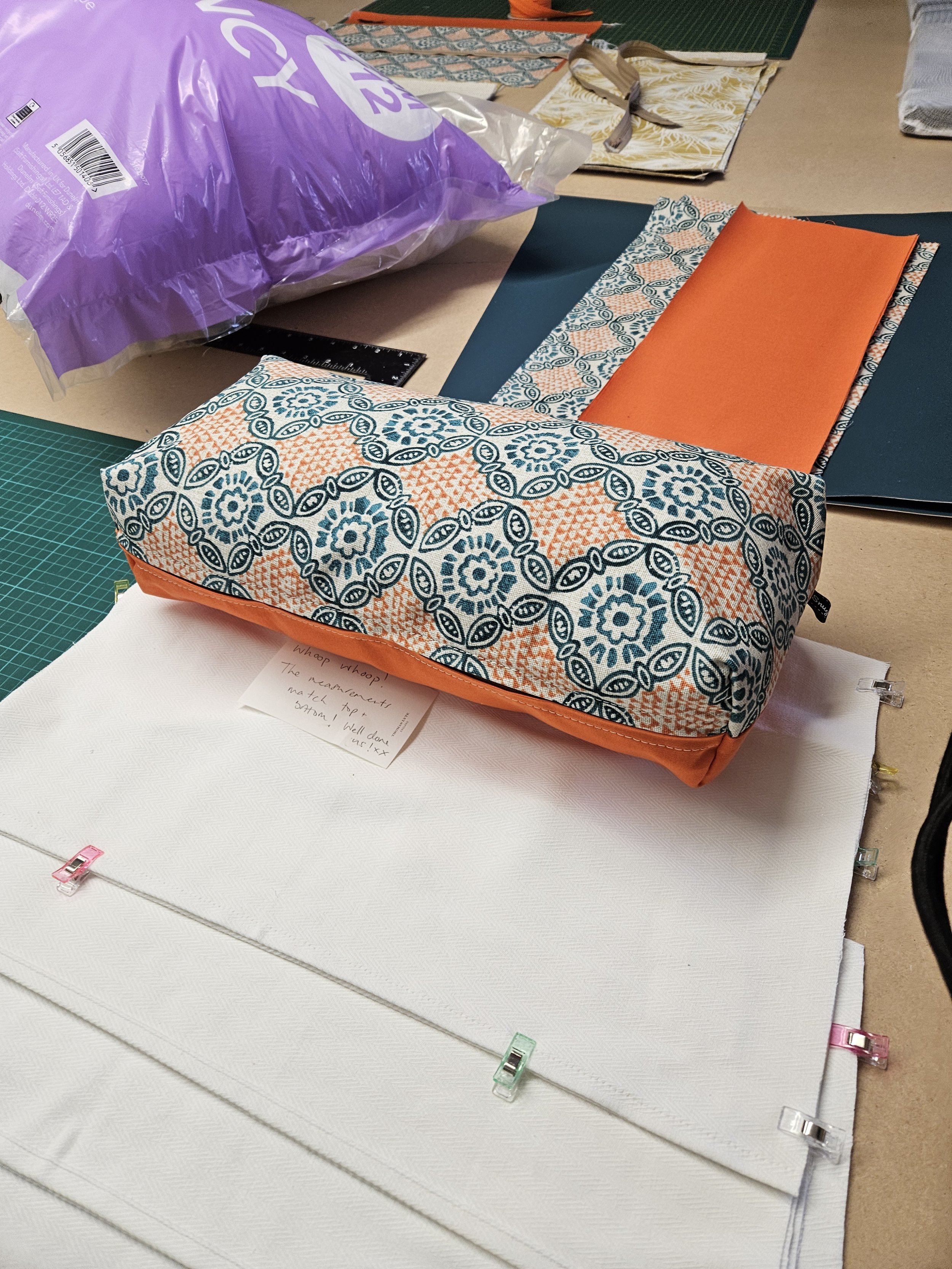 A desk or work table with various fabric pieces, notebooks, and a large purple plastic bag. A patterned fabric with floral and geometric designs in orange, blue, and white covers part of the table, with a solid orange fabric overlay. Several small cl