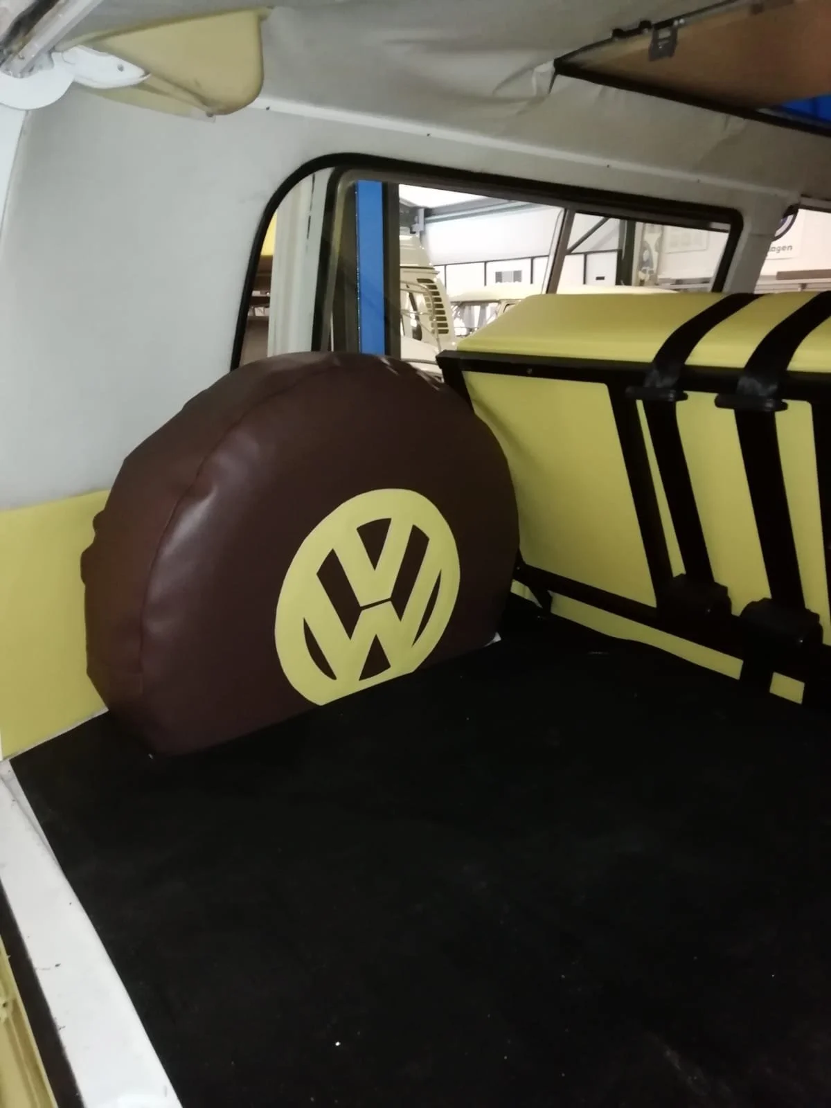 A brown cushion with a Volkswagen logo rests against the wall inside a vehicle, next to a yellow container and a music stand.