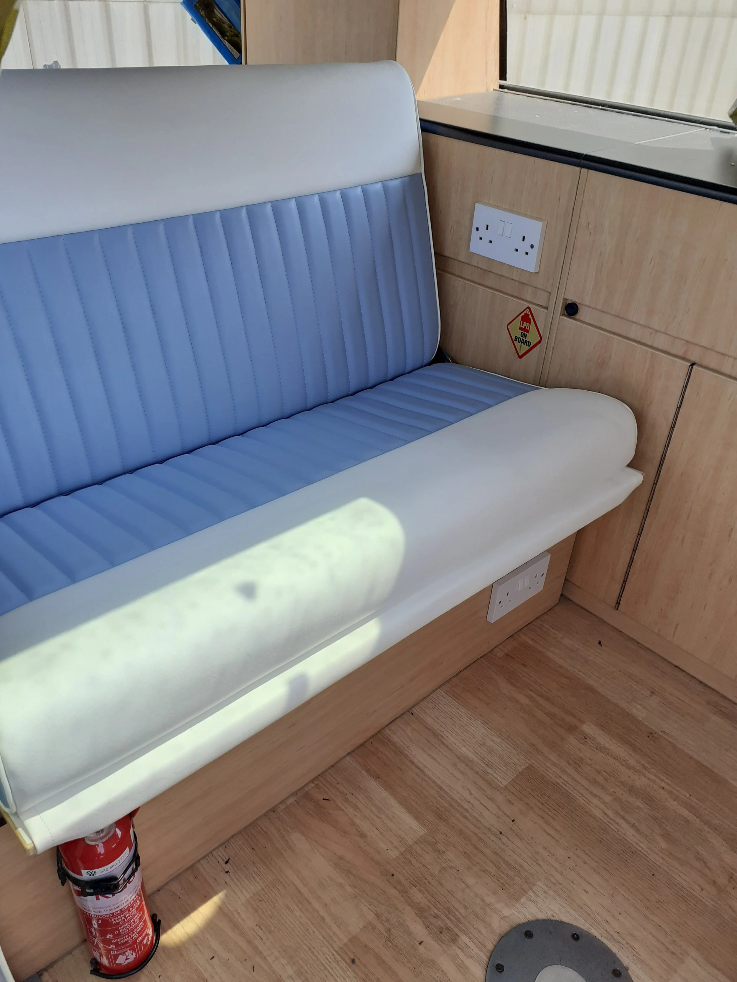 Blue and white vinyl bench seat next to electrical outlets and a warning sign on wooden panel, with a fire extinguisher at the bottom left corner, on a wood floor.