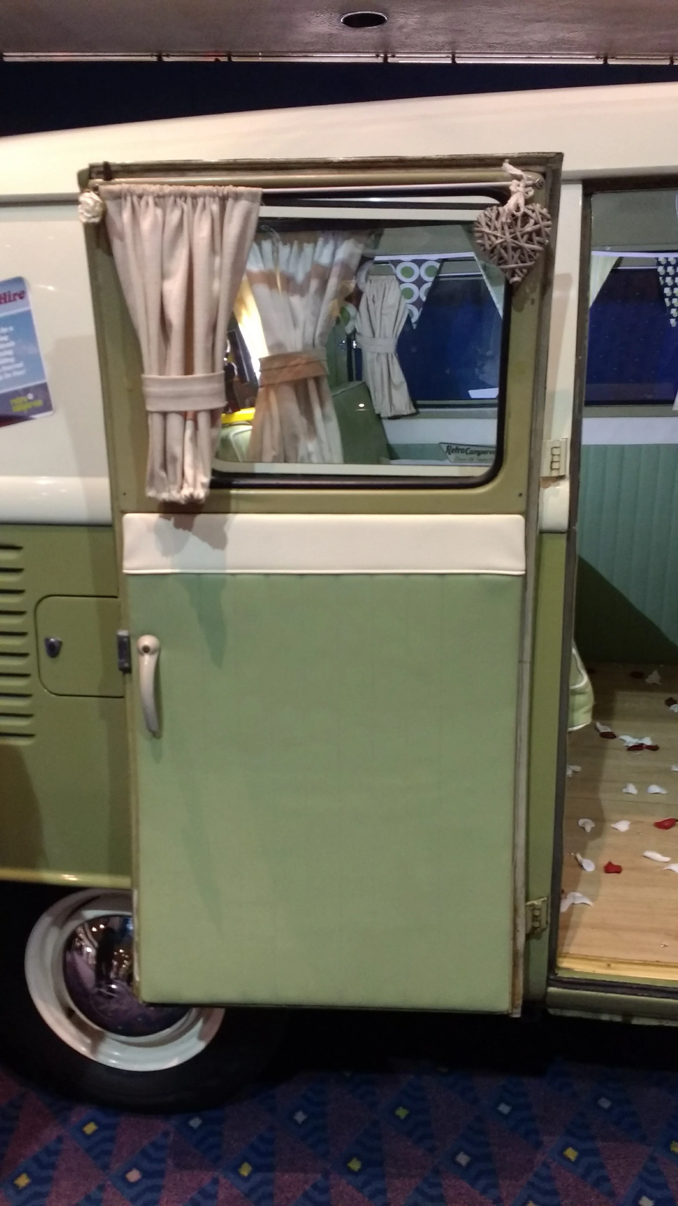 The photo shows a vintage green and white camper or trailer with curtains on the window, parked on a patterned carpeted floor, with scattered white and red paper decorations.