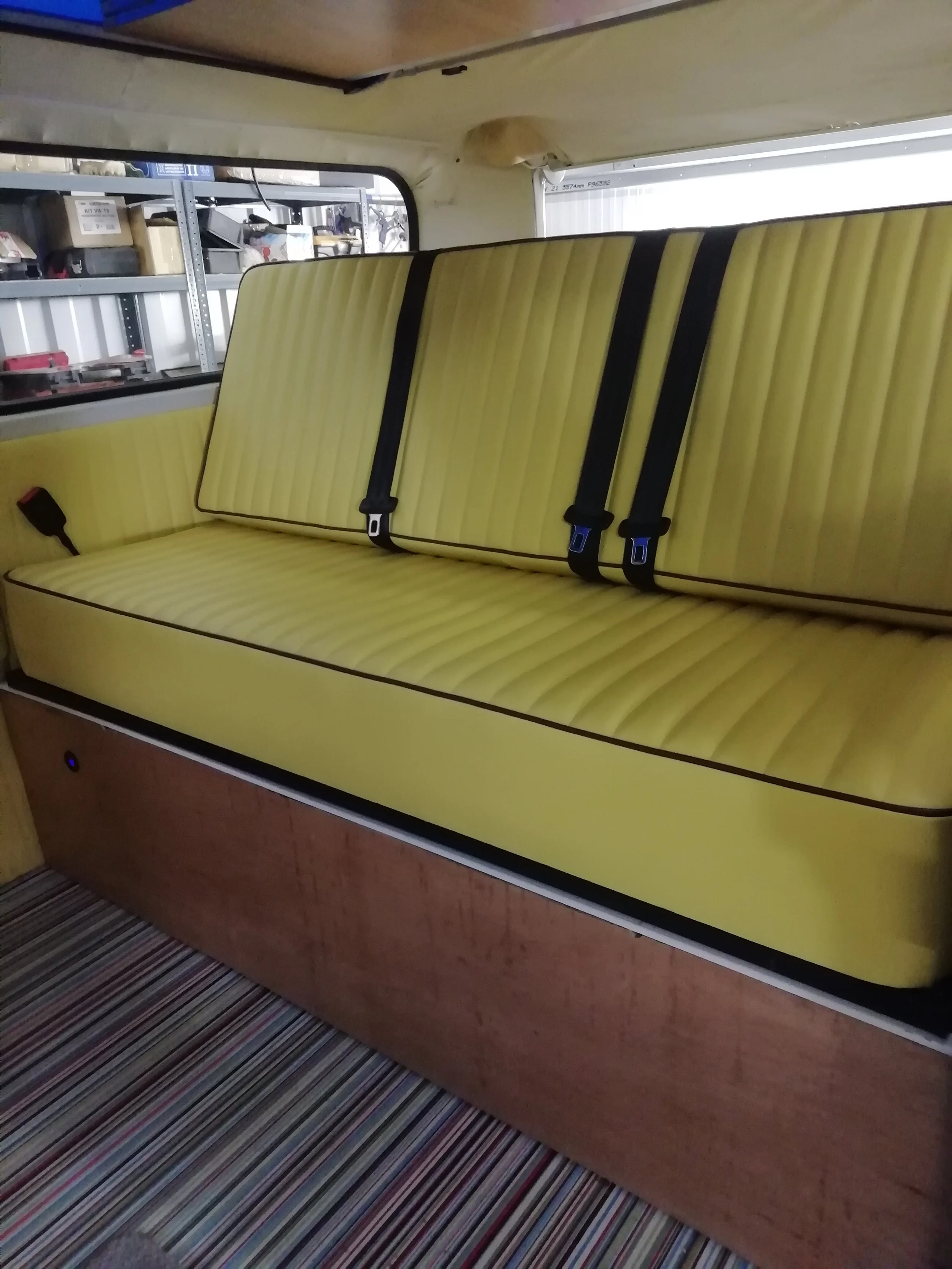 Yellow cushioned seat with black seatbelt straps, installed inside an aircraft, with a multicolored striped carpet on the floor.