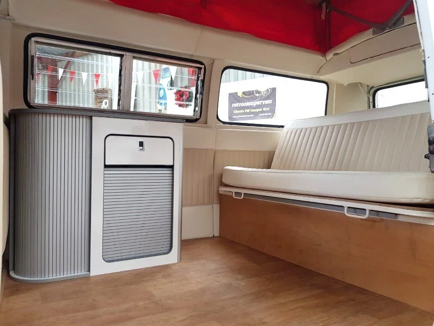 Interior of a vintage van with cream-colored seating, a small white cabinet, and wooden flooring, with windows showing exterior decorations.