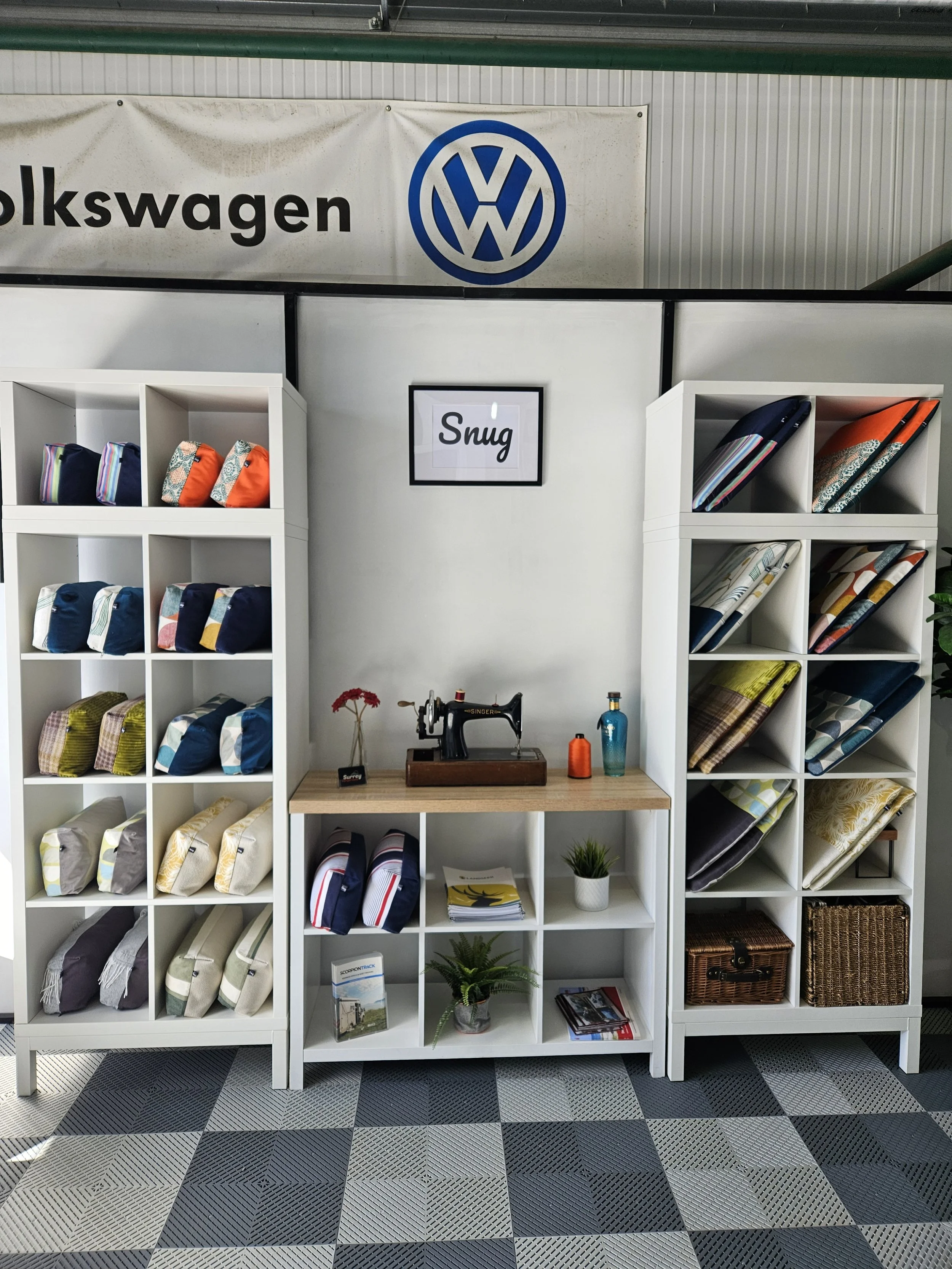 Display of fabric rolls and bags on white shelves at an interior space with a sign reading "Snug" and a sewing machine on a wooden table.