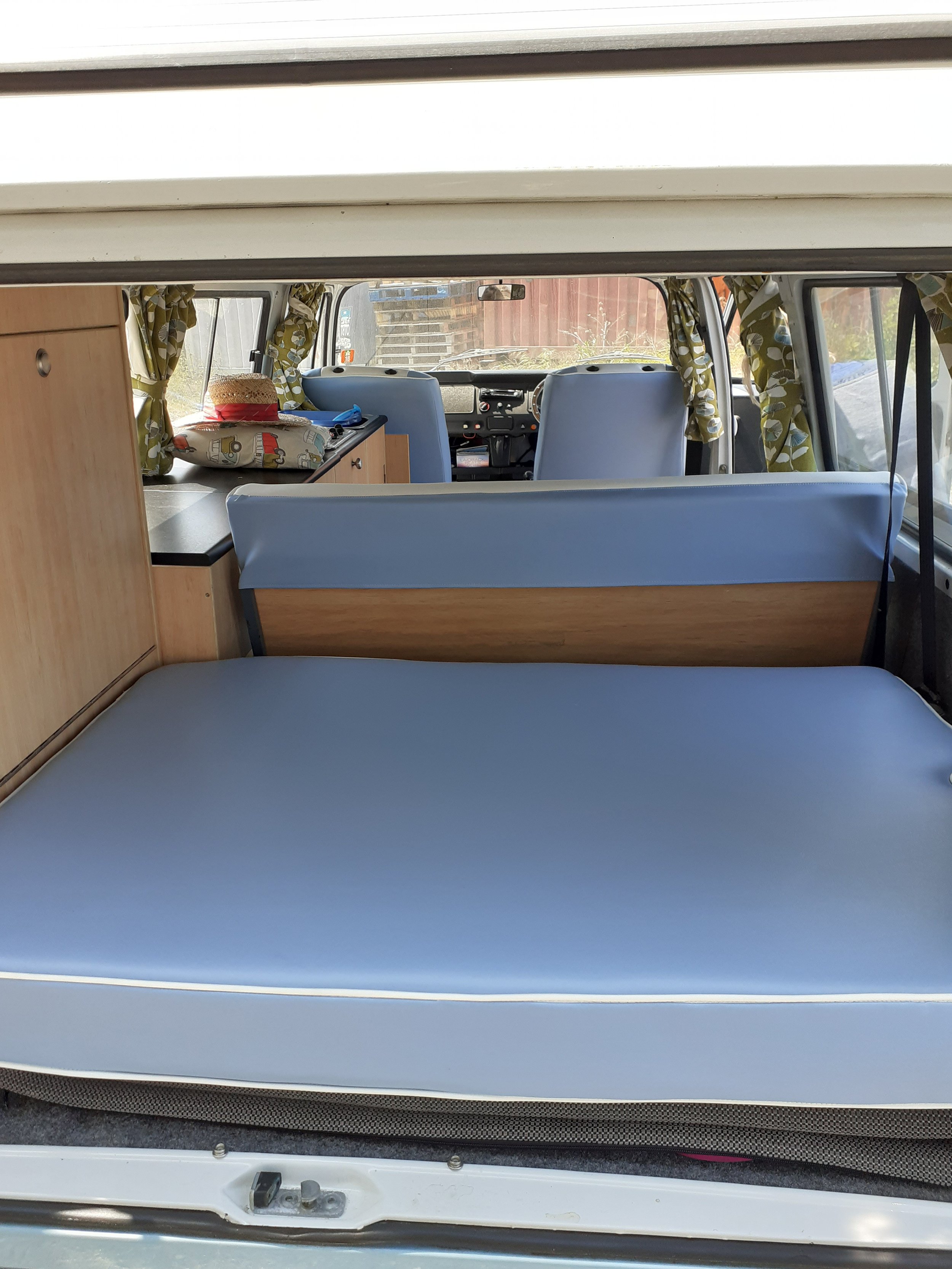 Inside view of a camper van showing the seating area and front cabin with curtain-covered windows, a hat, and a pillow on the seat.