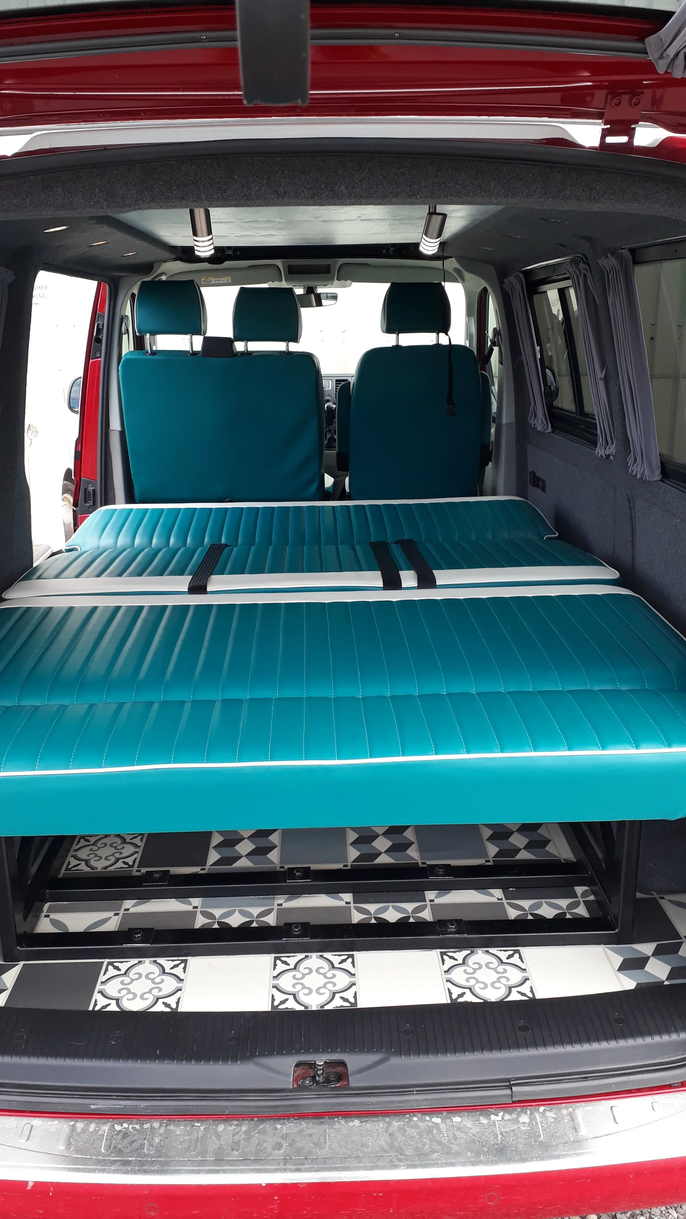 The interior of a red van, shown from the rear with the back door open, featuring two front seats, a folded mattress or cushion, and patterned floor tiles.