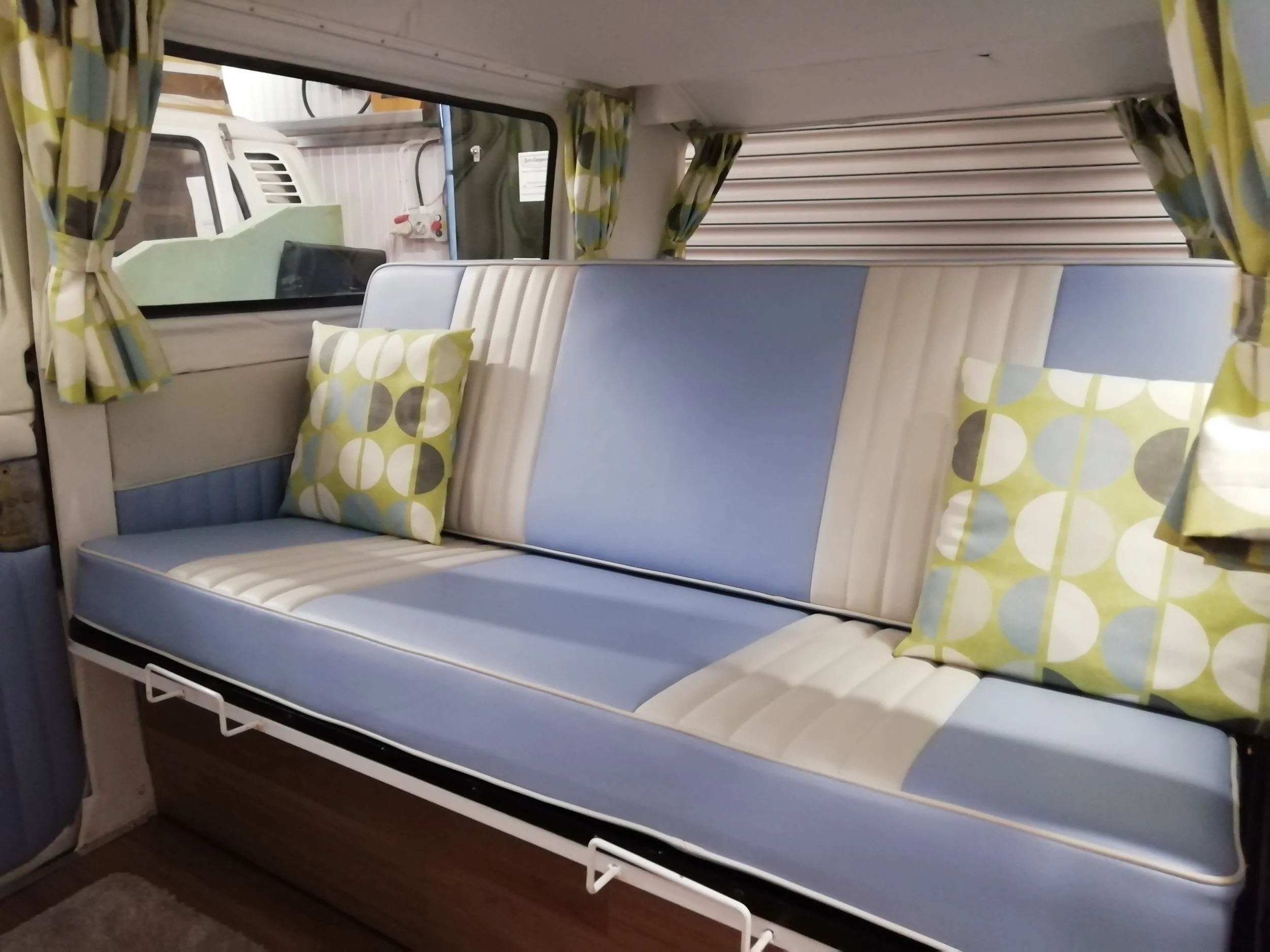 Interior of a camper with two beds, one above the other, featuring blue and beige striped mattresses and pillows with a green and beige circular pattern. The camper’s window is opened with green and beige patterned curtains that match the pillows. Ou