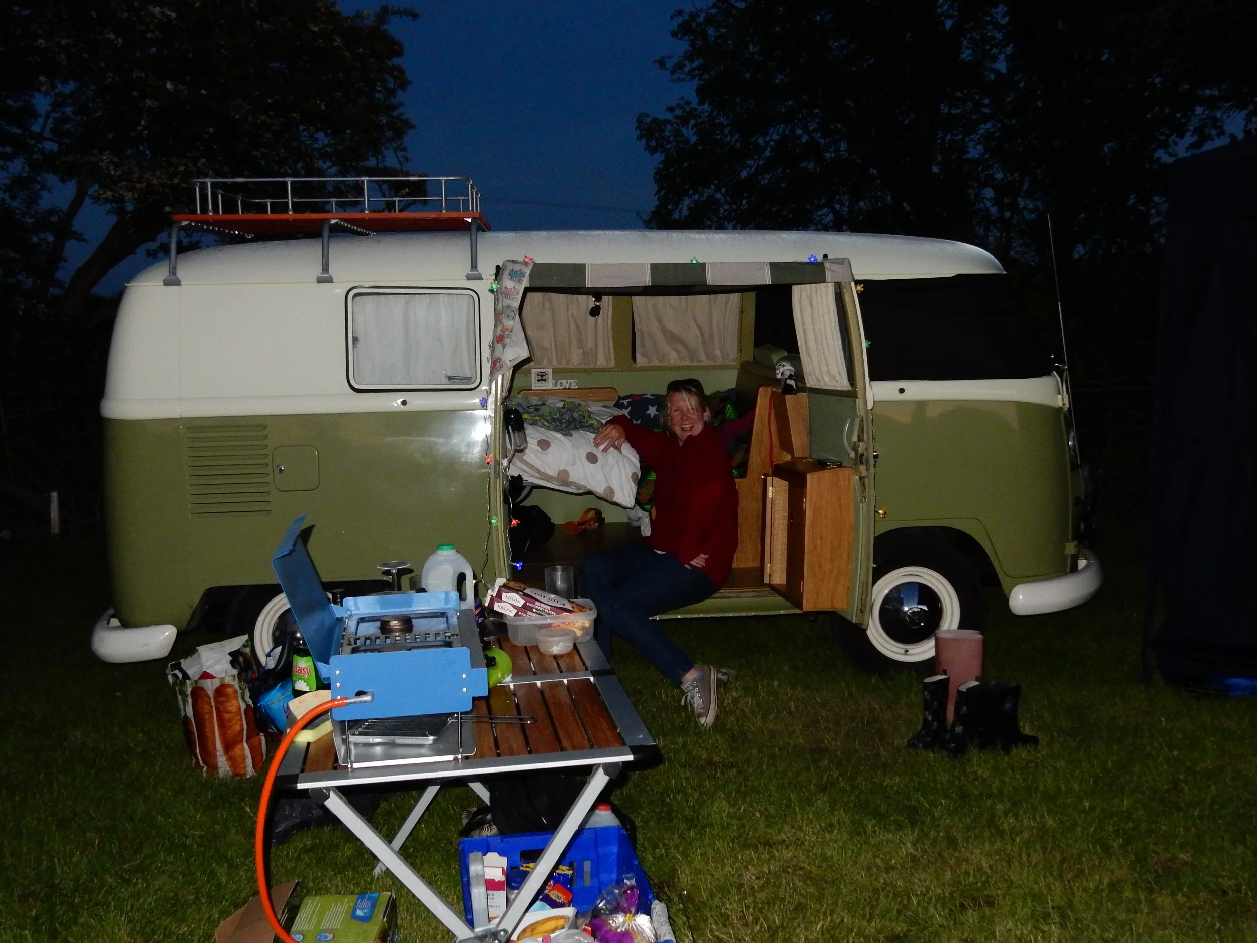 A woman with blonde hair smiling and lying inside a vintage camper trailer, which is parked outdoors at night. The camper has a window with curtains, and the background shows dark trees and sky. The area in front of the camper is set up with various camping and cooking supplies, including a foldable table, a portable stove, food items, and utensils.
