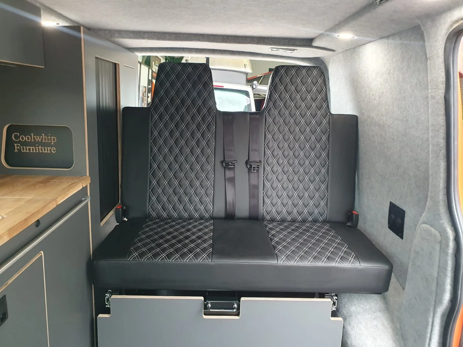 The interior of a van with a black two-seater bench seat with diamond-patterned upholstery. Behind the seat, there are windows and part of the vehicle's interior wall with a speaker and window controls.