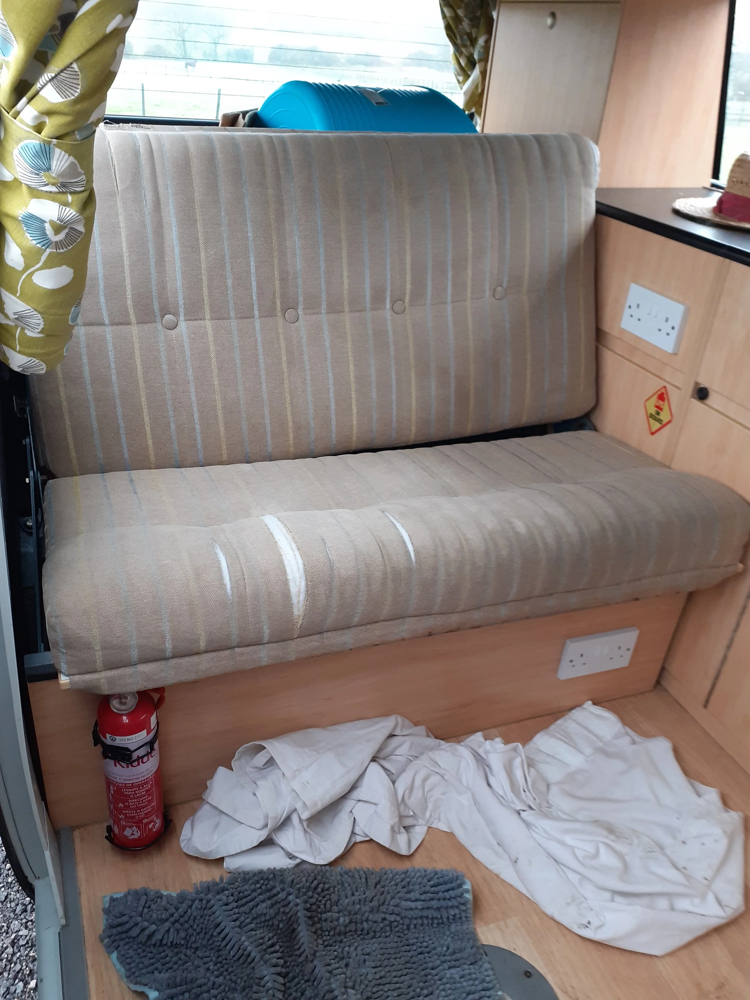 The image shows the interior of a vehicle or possibly a mobile home, with a beige striped upholstered bench or bed, crumpled white cloth or paper on the floor, a fire extinguisher next to it, a gray textured mat, electrical outlets on the wooden pane