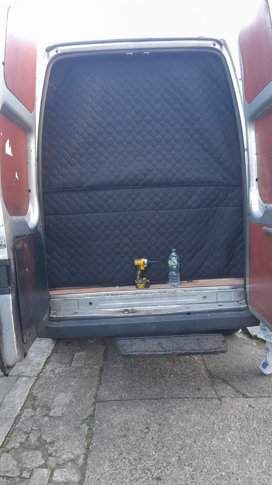 Empty vehicle cargo area with black quilted lining, a yellow cordless drill, and a water bottle on the floor, viewed from outside.