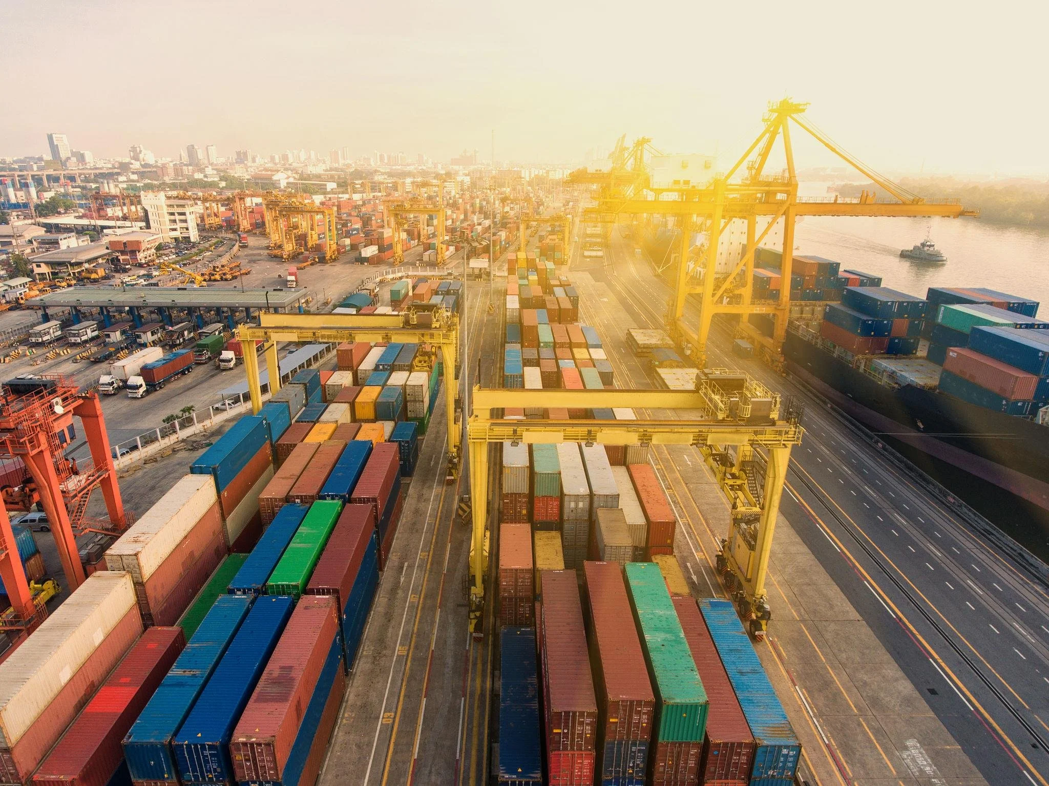 Aerial view of a busy port with numerous shipping containers, large yellow cranes, trucks, and a cargo ship, set against a city skyline with high-rise buildings in the background, under a hazy sky.
