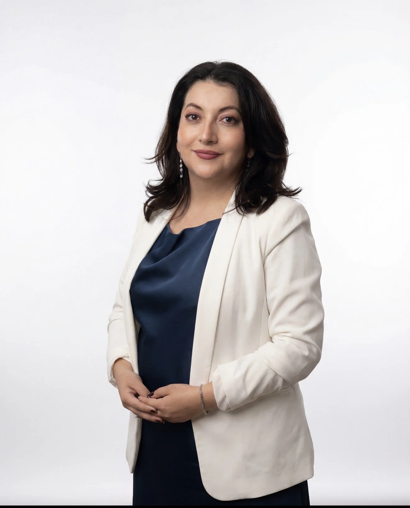 Professional woman with dark hair, wearing a white blazer over a dark blue top, standing against a plain white background.