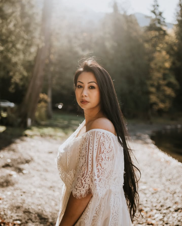 A woman with long dark hair standing outdoors near a lake or river in a forest. She is wearing a white lace off-shoulder dress, looking at the camera with a serious expression. The background includes trees and sunlight filtering through the leaves.