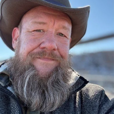 Close-up of a man with a beard wearing a cowboy hat and outdoor jacket, smiling at the camera with a slightly blurred background and blue sky.