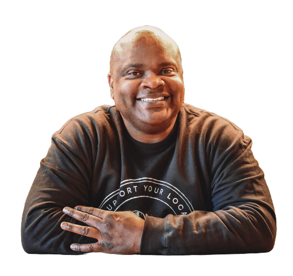 A smiling black man with a shaved head wearing a black long-sleeve shirt with a circular design, sitting with arms crossed and looking at the camera.