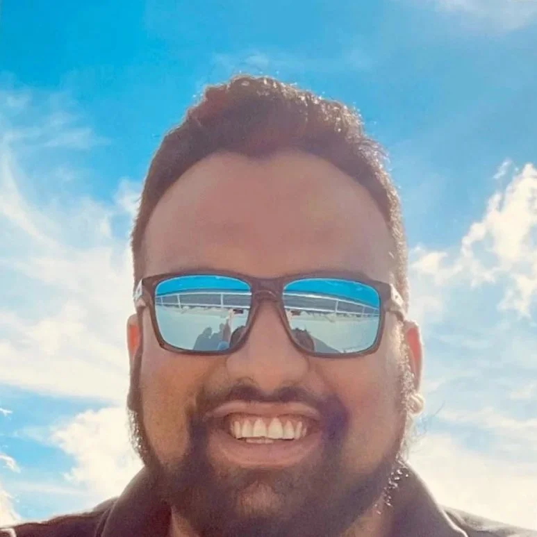 A man with curly dark hair, beard, and sunglasses smiling outdoors with blue sky and scattered clouds.