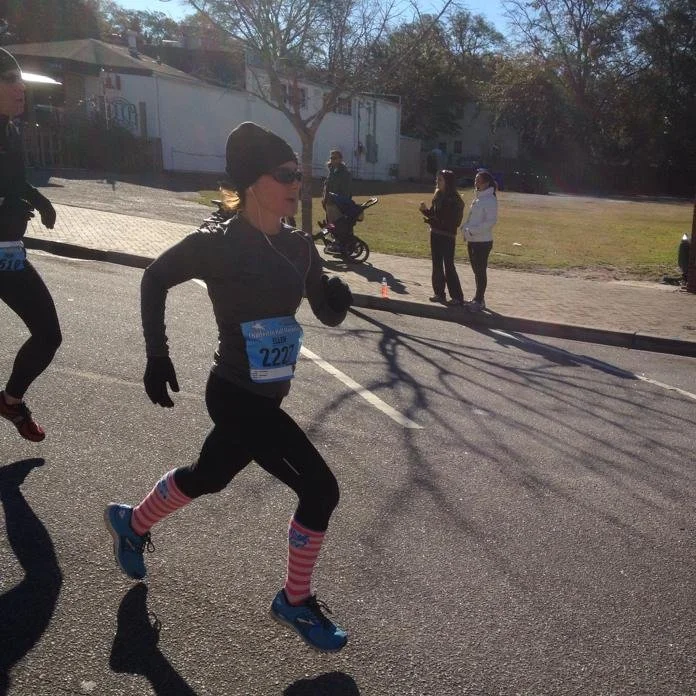 A woman running in a road race, wearing sunglasses, gloves, black athletic clothing, a race bib number 227, and colorful striped knee-high socks, with spectators in the background on a sunny day.