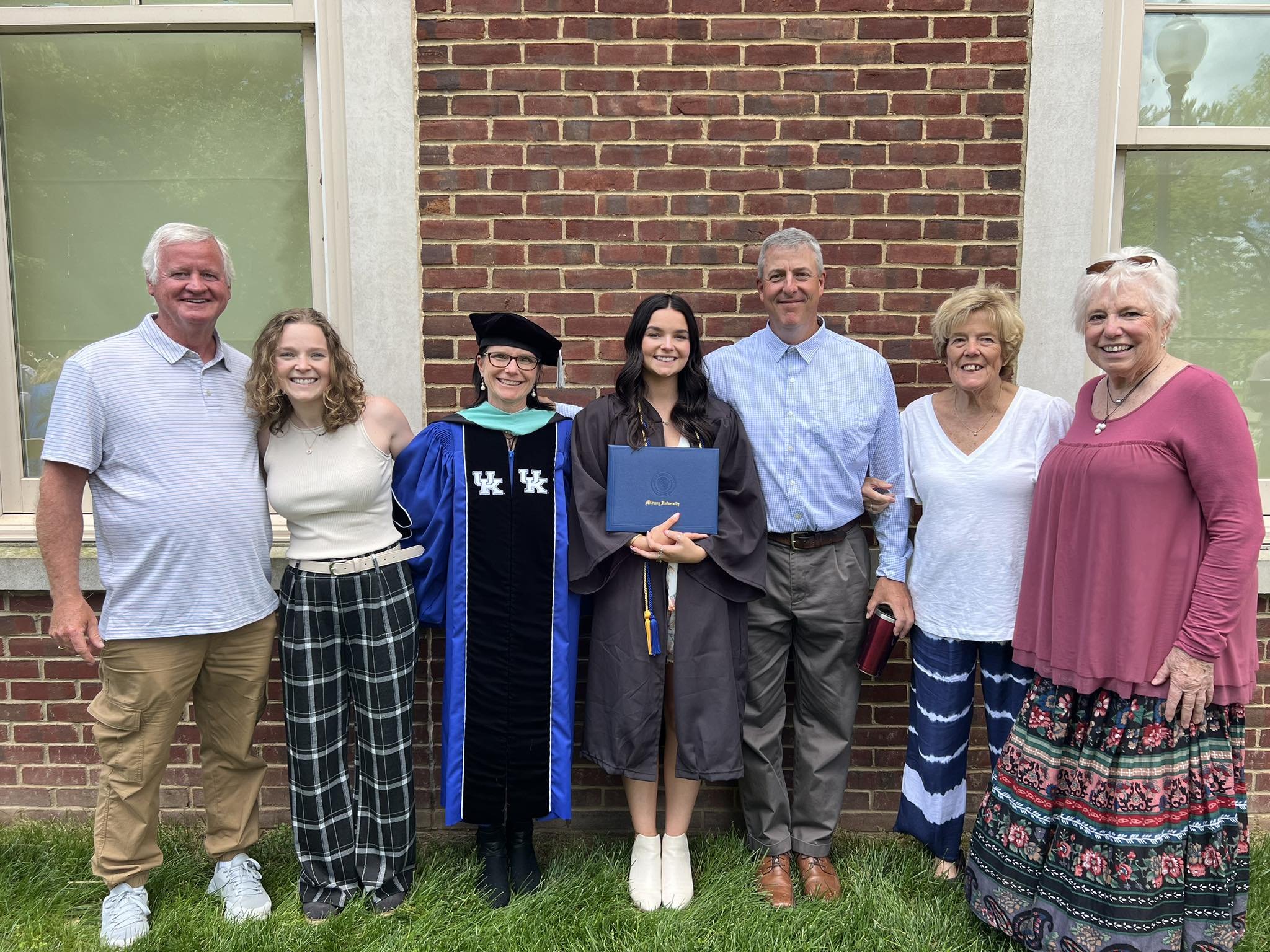 A young woman in a graduation gown and cap holding a diploma, standing with several family members in front of a brick wall.