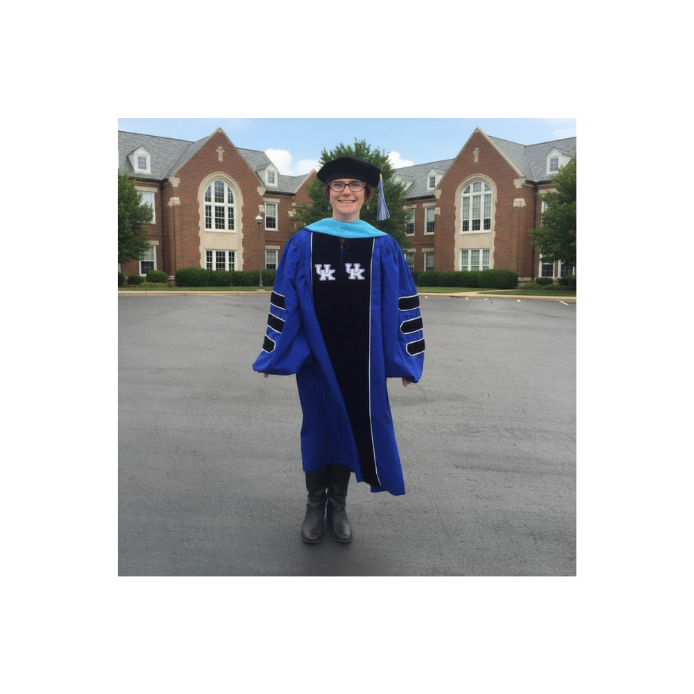 Person in graduation cap and gown standing on a paved area in front of a large brick building with trees.