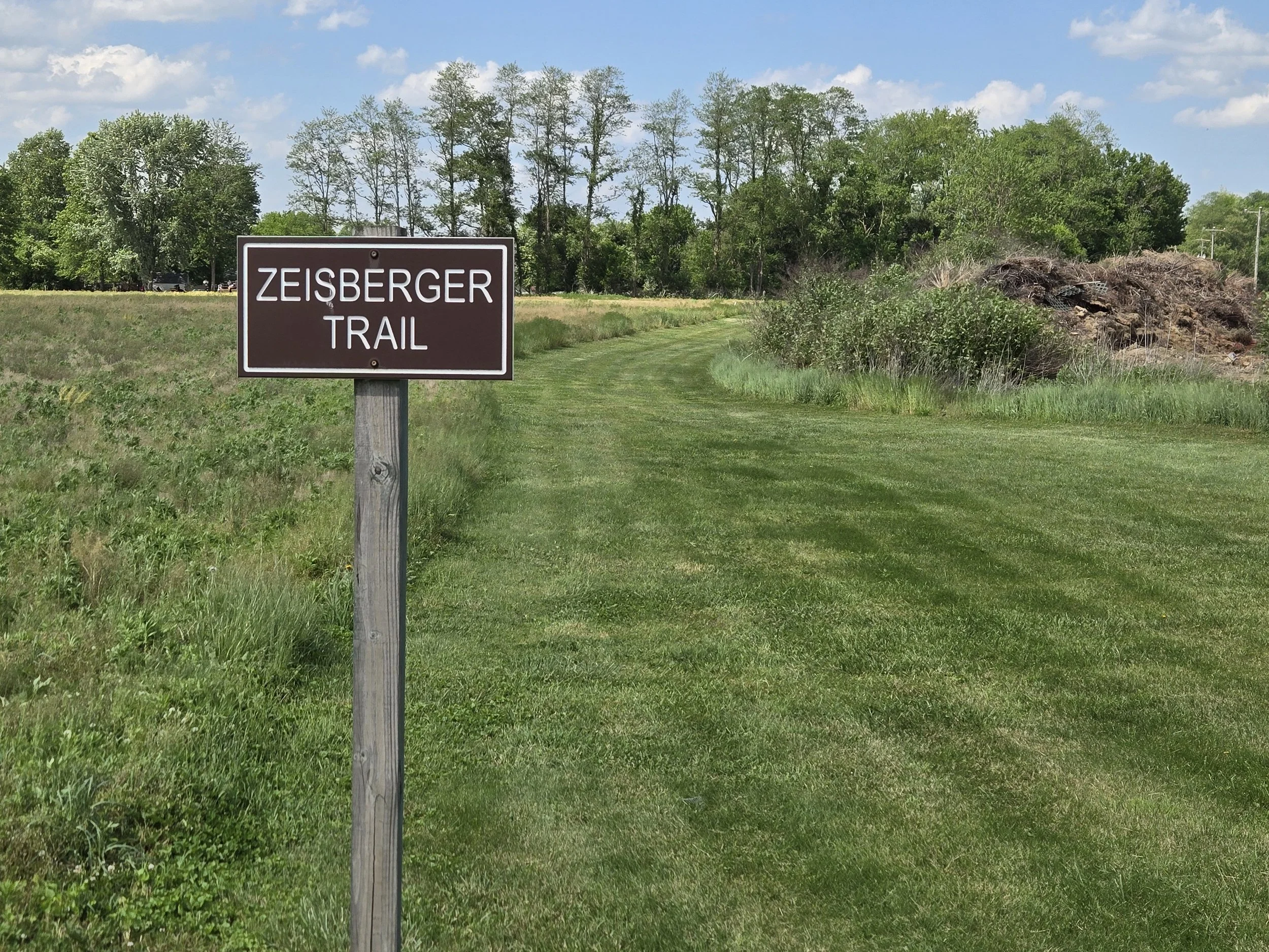 A park trail sign reading 'Zeisberger Trail' placed on a grassy area with trees in the background under a partly cloudy sky.