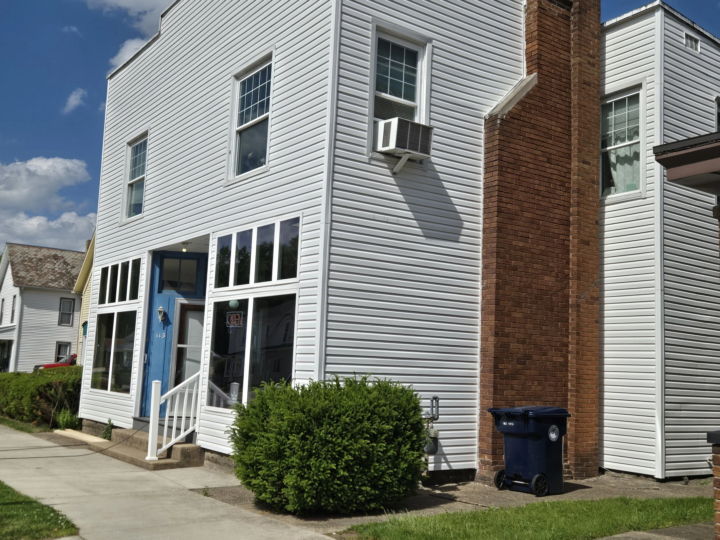 A three-story white house with a brick chimney and an air conditioning unit in one window. The house has a small front porch with white railings, a blue door with the number 113, and large windows. There is a neatly trimmed bush in front, a sidewalk, and a trash bin to the side. The sky is blue with some clouds.