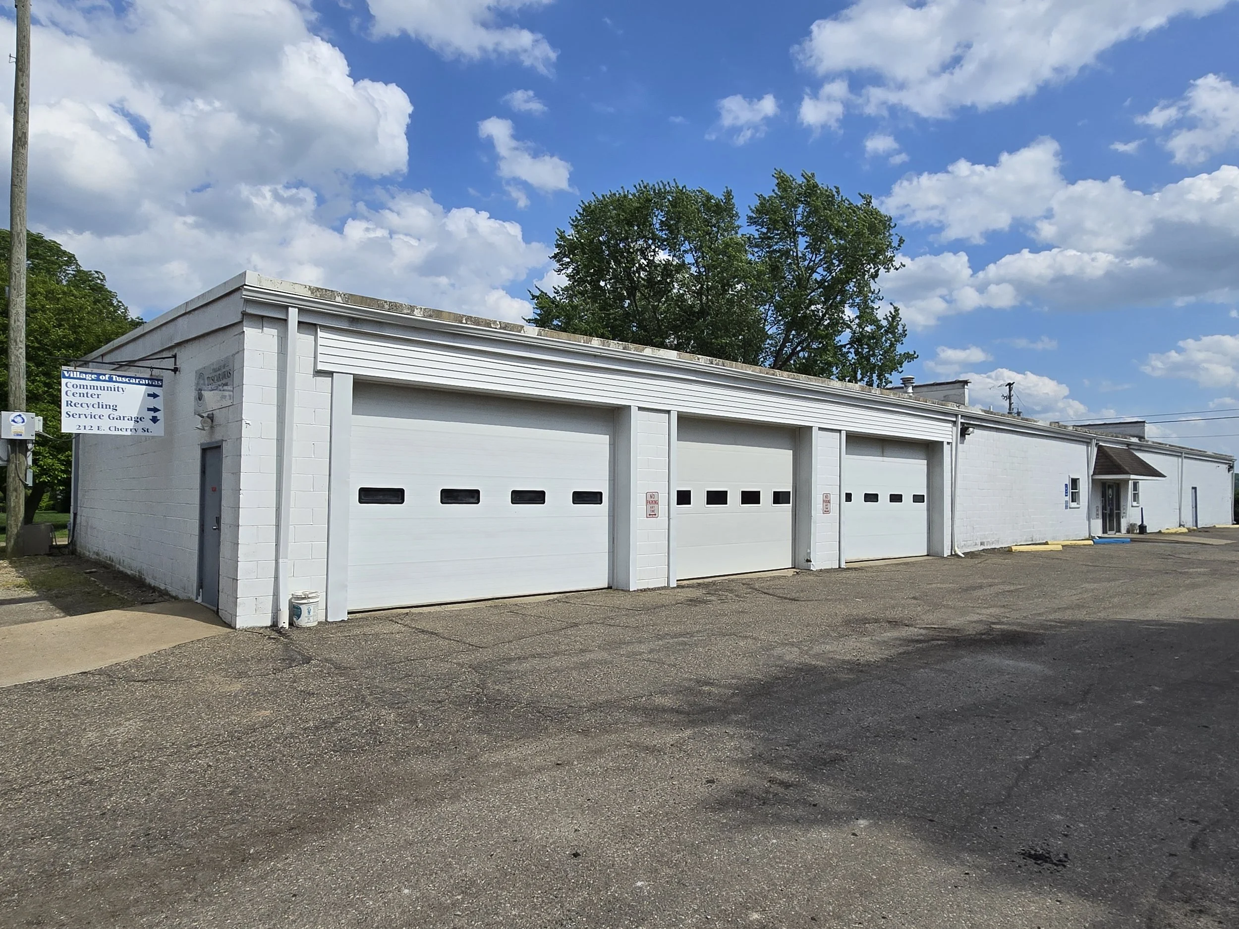 A white building with three garage doors and a sign indicating it is a community center and recycling service garage on Cherry Street. The building is adjacent to a parking lot with a rough asphalt surface. There are some trees in the background, a utility pole on the left, and a partly cloudy sky overhead.