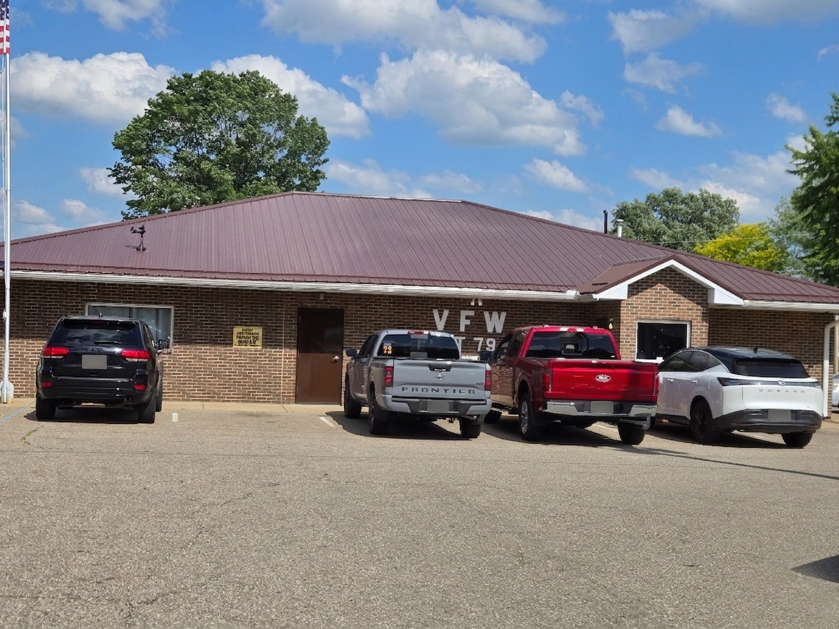 A brick building with a maroon metal roof and the letters "V F W" and the number "79" painted on the front, with four vehicles parked in front under a partly cloudy sky.