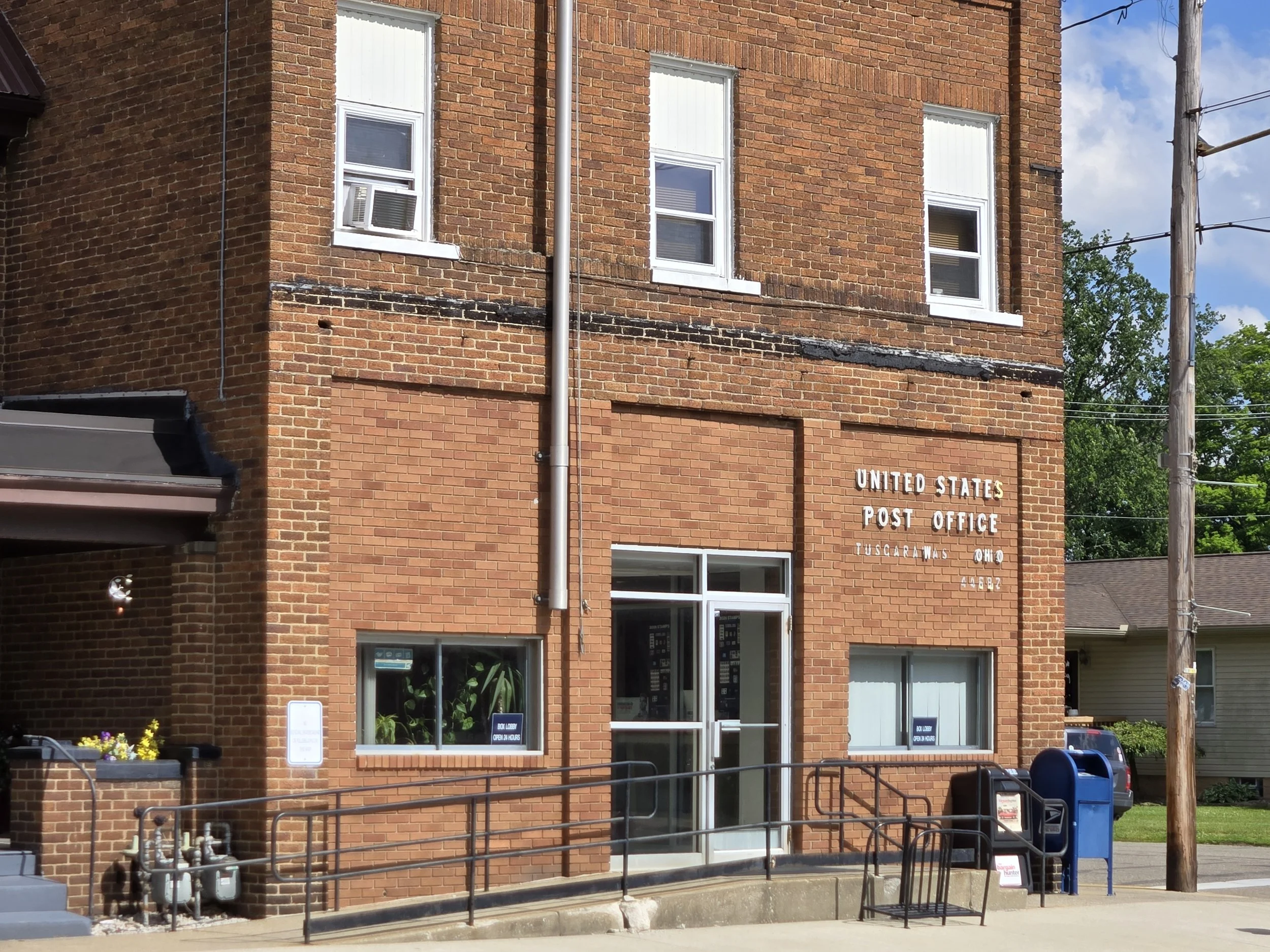 Front view of a brick building with a sign that reads 'United States Post Office' in Tuscarawas, Ohio. The building has multiple windows, a glass door, a blue mailbox outside, and a small flower arrangement near the entrance. There are utility poles and power lines visible along with some trees and a partly cloudy sky.