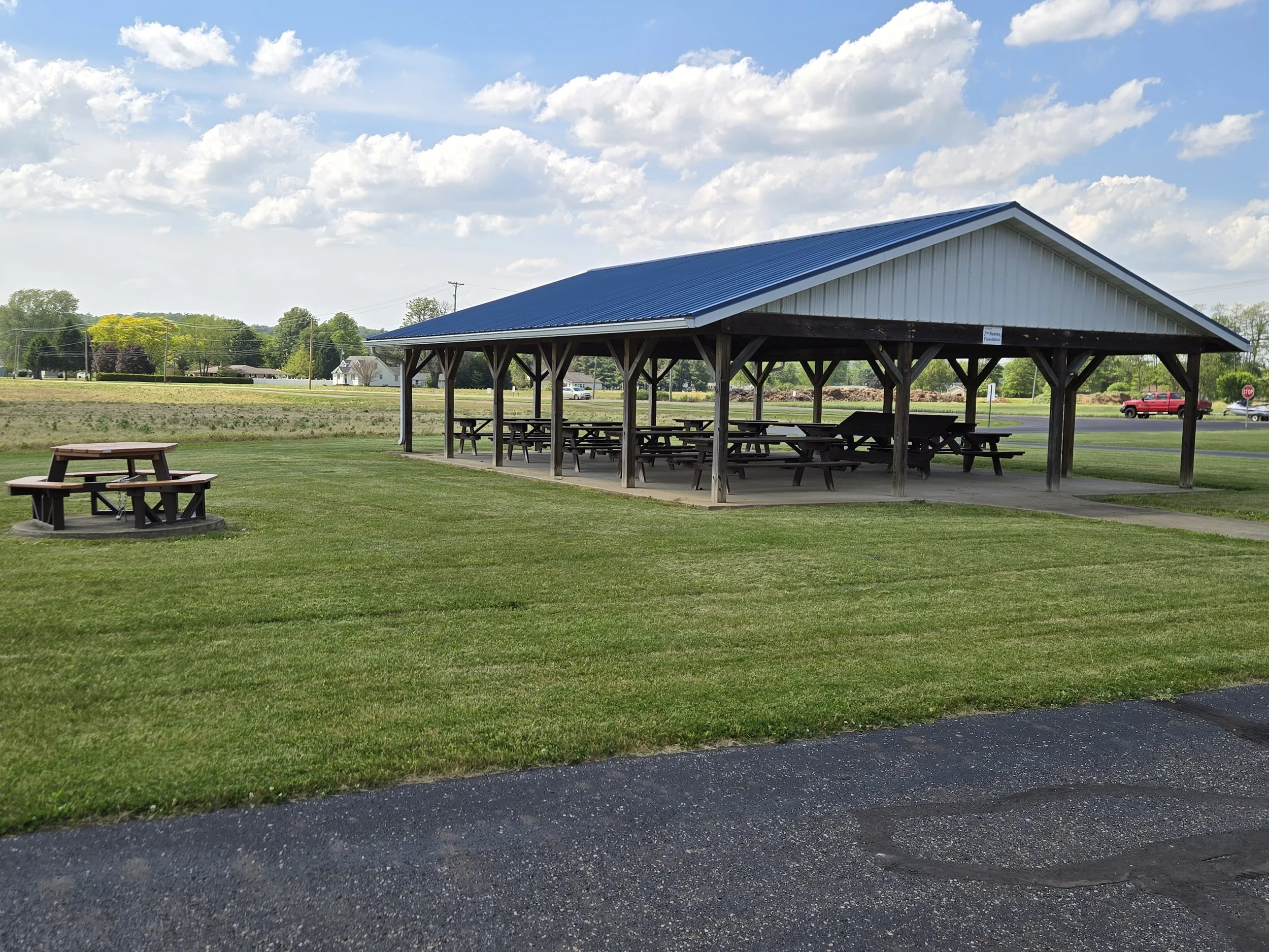 A park shelter with a blue metal roof, surrounded by green grass and trees under a partly cloudy sky.