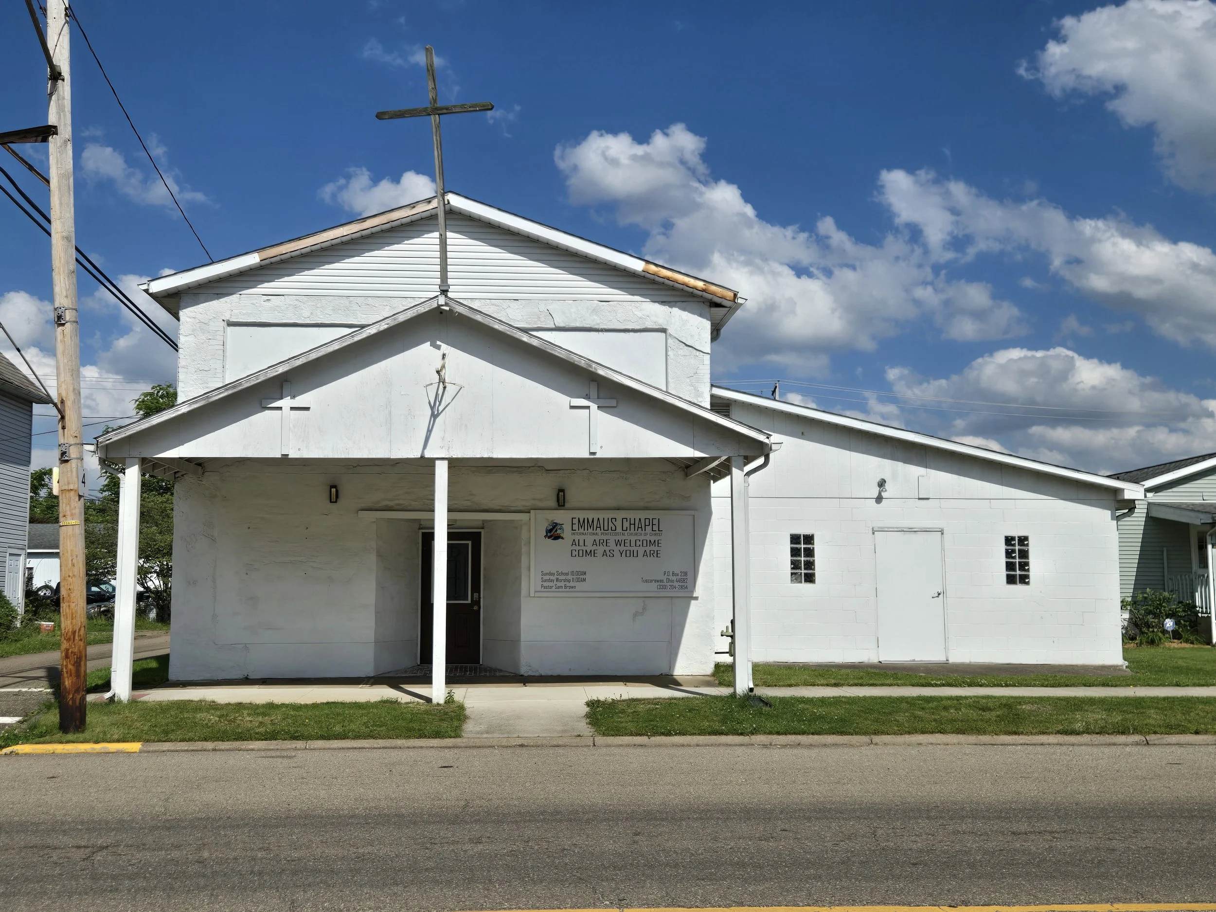 Front view of Emmaus Chapel, a white building with a small porch, cross on top, and a sign that reads 'Emmaus Chapel. All are welcome. Come as you are.'