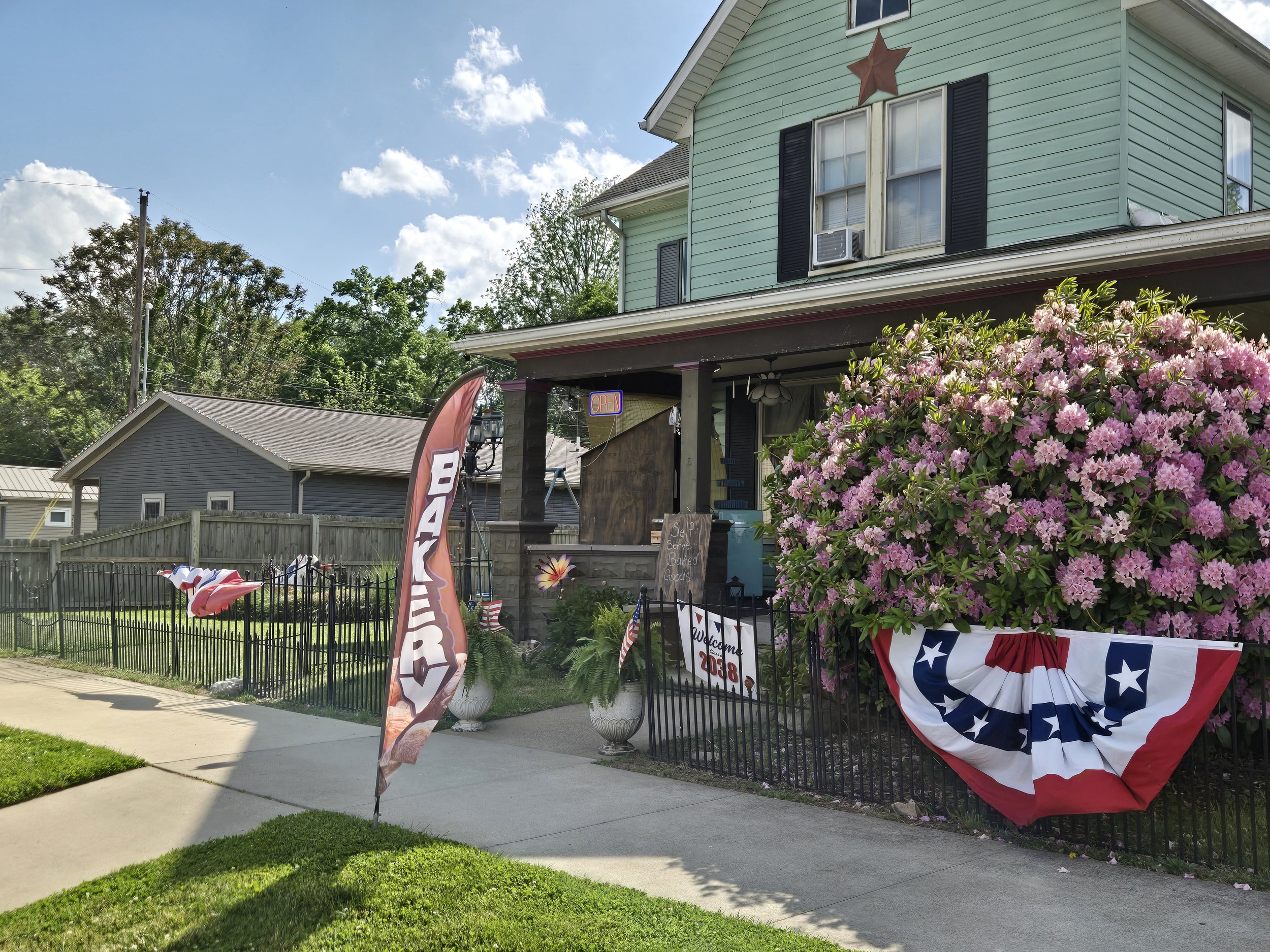 A two-story light green house decorated with patriotic red, white, and blue banners and flowers, with an 'OPEN' sign hanging from the porch, and pink flowering bushes in front.