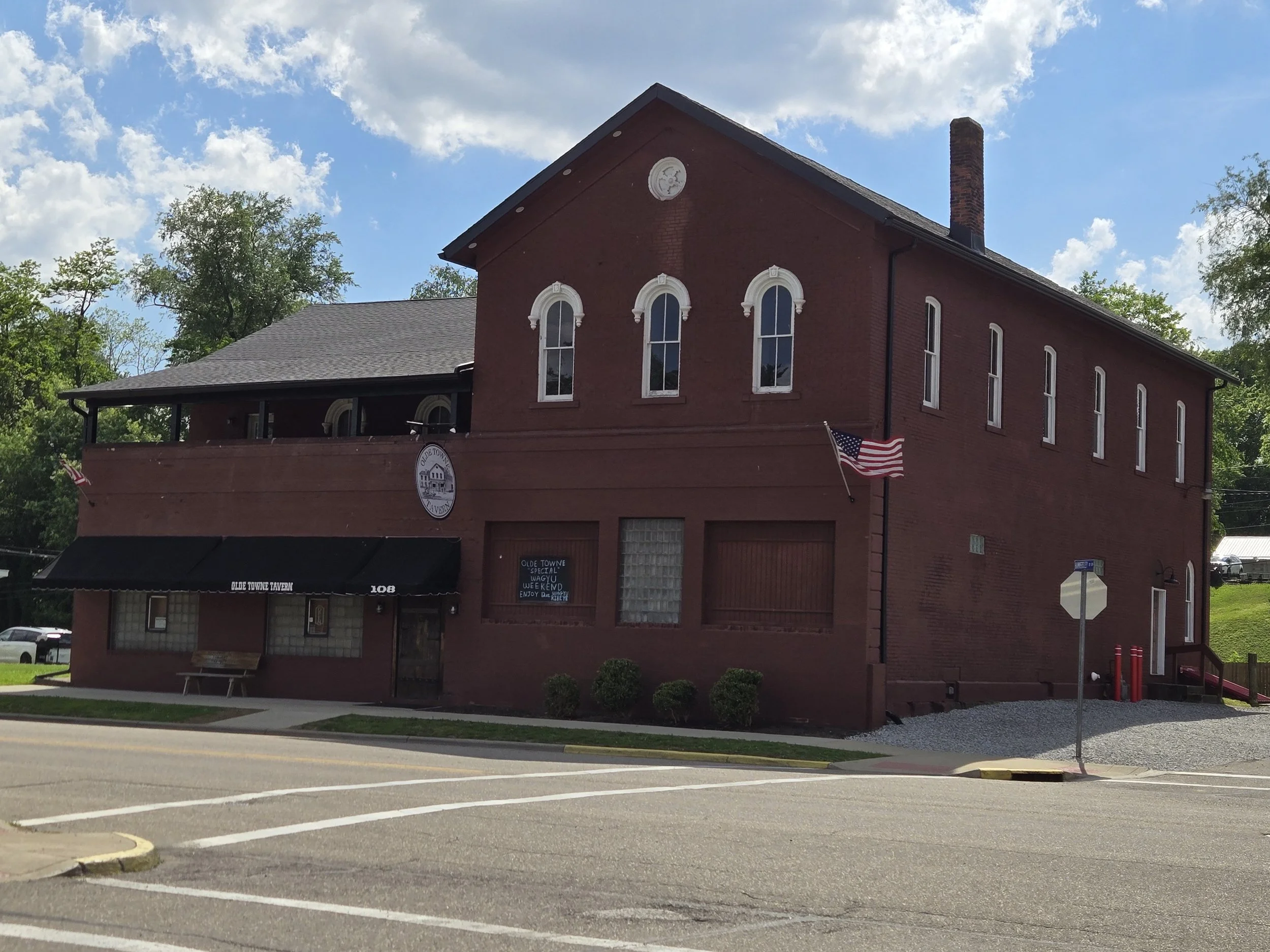 A two-story red brick building with arched windows and a chimney, featuring a sign for Olde Towne Tavern, with a sidewalk, an American flag, and trees in the background, under a partly cloudy sky.