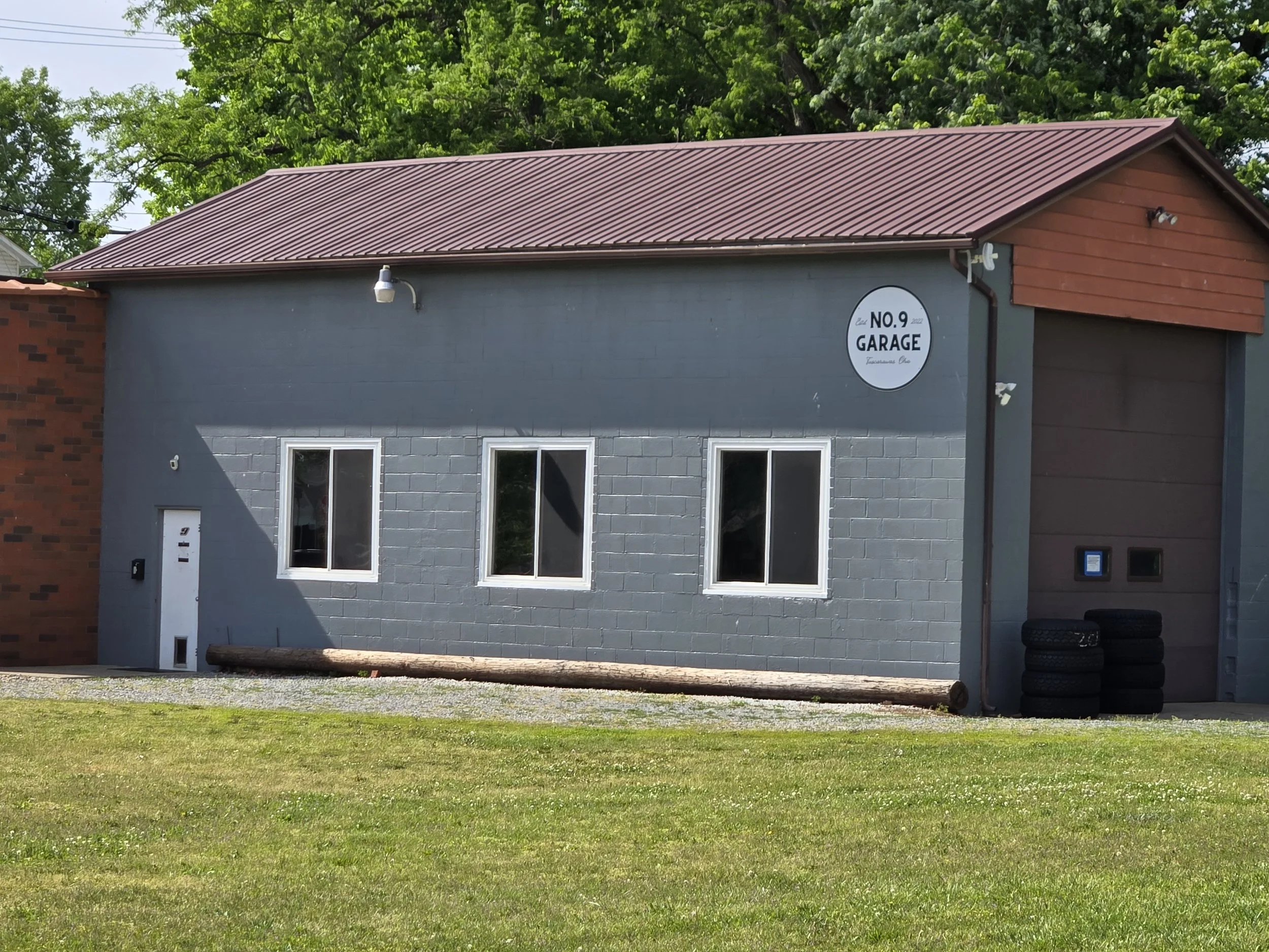 A gray building with a red metal roof, three windows, and a sign that reads 'No. 9 Garage'. There are two outdoor lights and a pile of tires in front of the building on the right side. A log is lying on the grass in front of the building, and tall green trees are in the background.