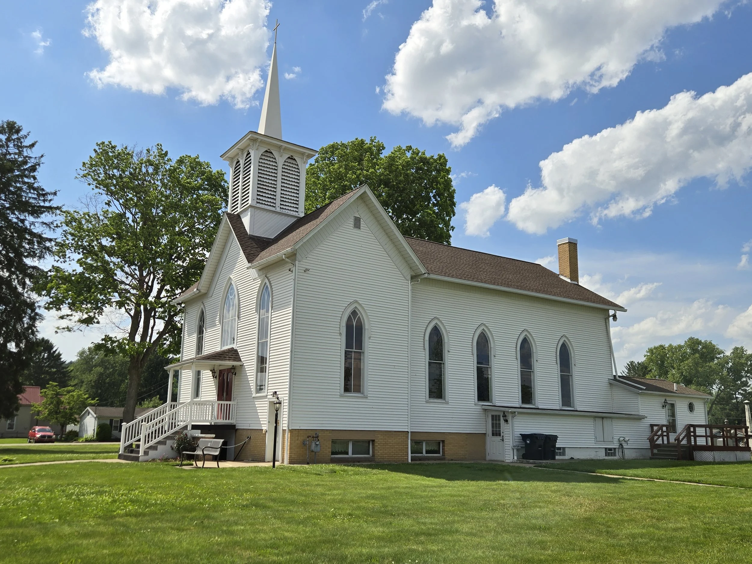 A white church with tall steeple, multiple arched windows, surrounded by green grass and trees, under a partly cloudy blue sky.
