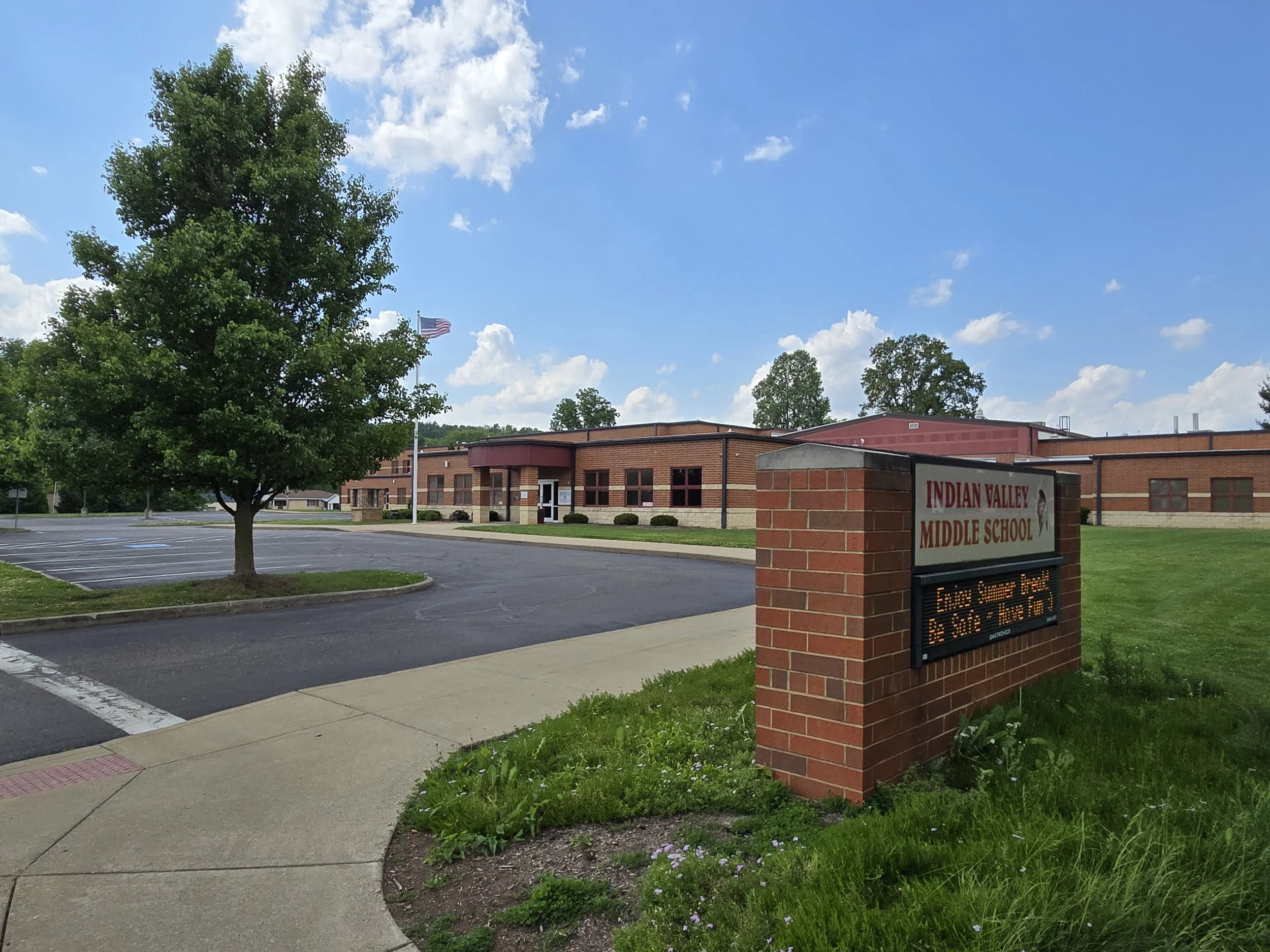 Exterior view of Indian Valley Middle School with a tree in the foreground, a blue sky with scattered clouds, and a sign displaying the school's name.