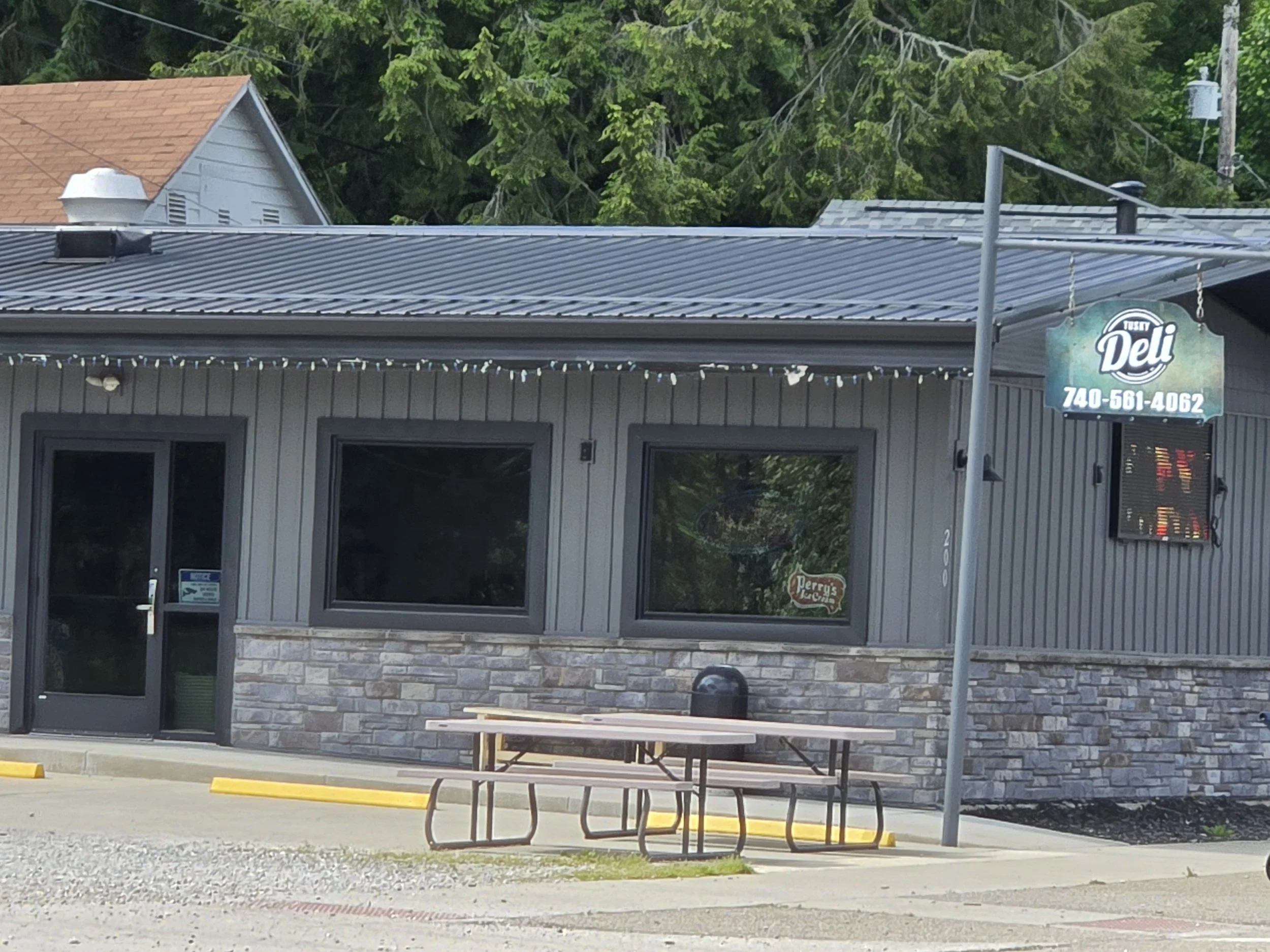 The exterior of a gray building with large windows and a door, featuring a stone facade at the bottom, and string lights hanging along the top of the windows. There is a roadside sign for a business called "Tasty Deli" with a phone number. In front of the building, there is a picnic table and parking spaces with yellow wheel stops. The background shows trees with green foliage and neighboring rooftops.