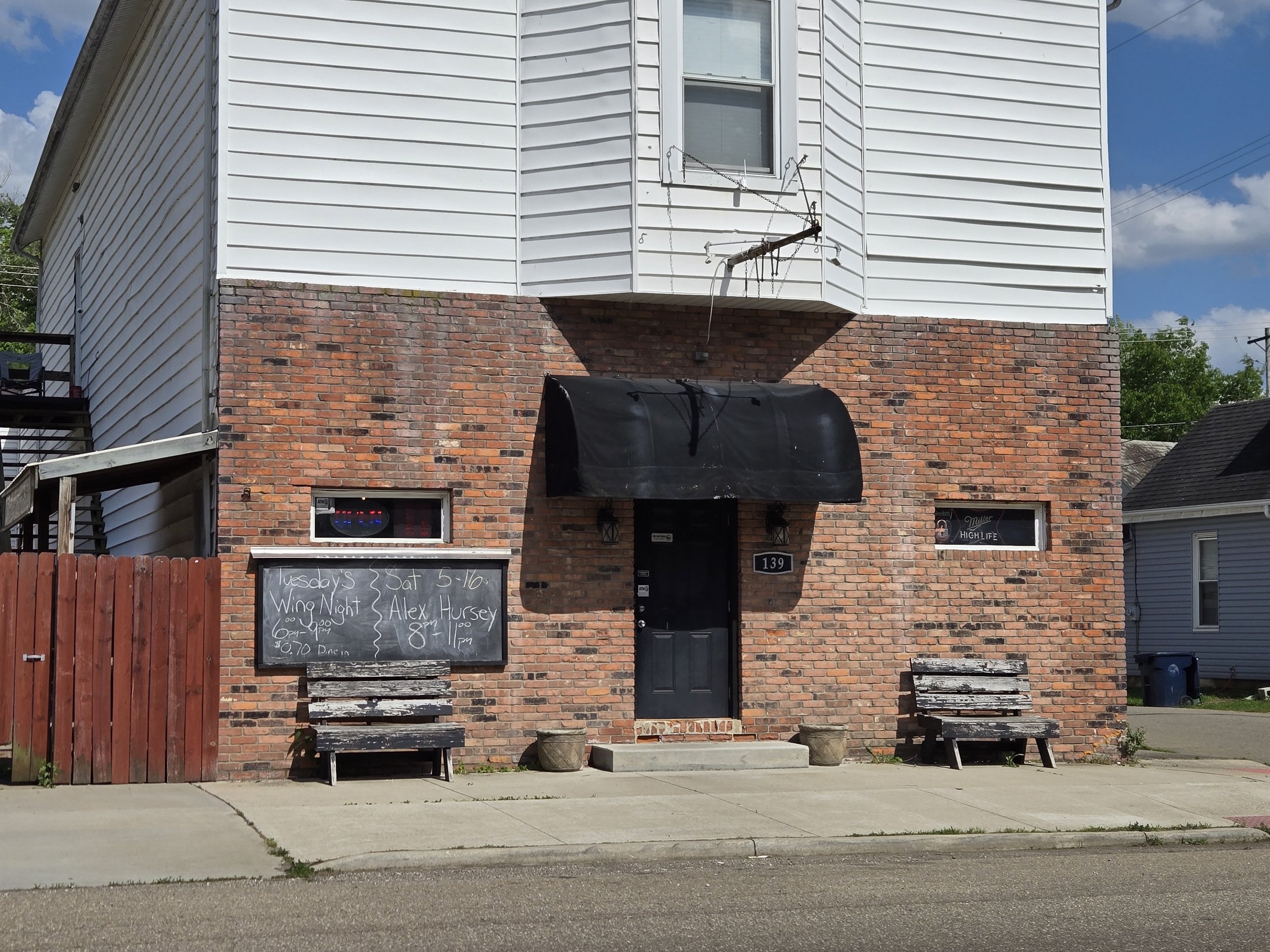 A brick building with a black door, two wooden benches, and a chalkboard sign advertising a Wing Night event. The sign indicates the event is on Tuesday and Saturday from 5-10 PM and features live music by Alex Hursey from 8-11 PM. There are two small windows on the building with some decorations, a fenced yard with a fire pit, and outdoor chairs visible.
