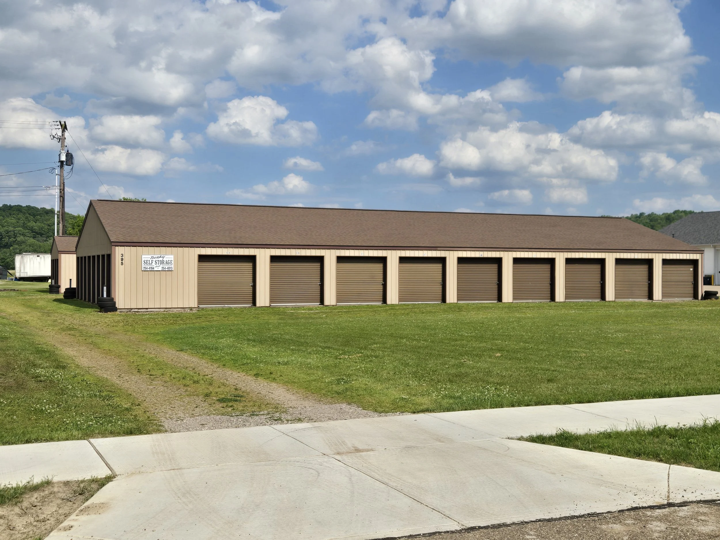 A storage building with a brown roof and beige exterior, situated on a grassy area with a sidewalk in front, under a partly cloudy sky.