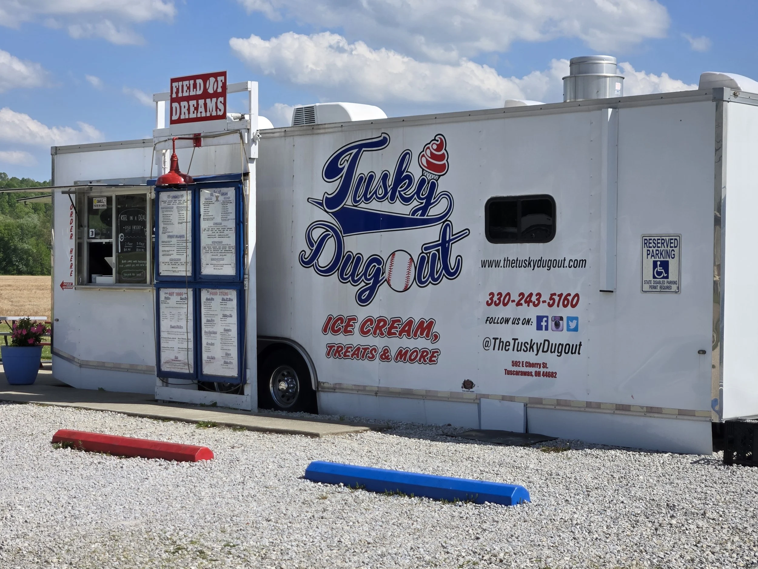 A white food truck called 'Tusky Dugout' offering ice cream, treats, and more, with a sign that reads 'FIELD OF DREAMS' on top, parked under a blue sky with scattered clouds.