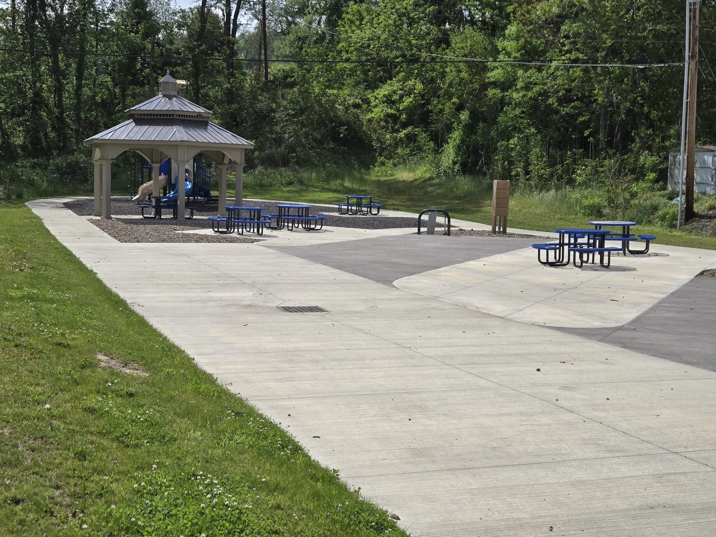 A playground area with a gazebo, blue picnic tables, and surrounding greenery, with trees in the background and a concrete surface.