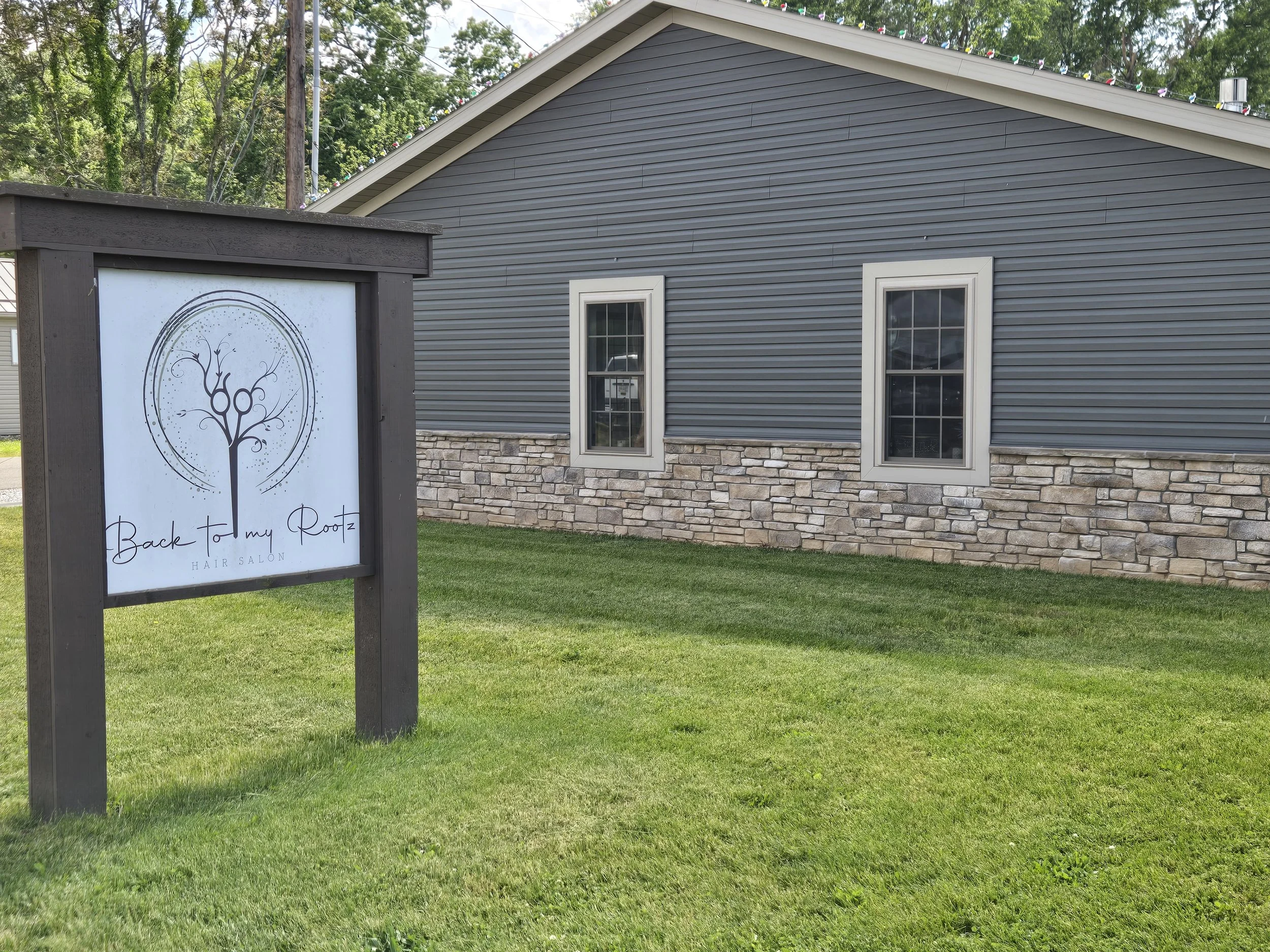 A gray, two-story building with stone accents at the bottom, two windows on the side, and a sloped roof with colorful string lights along the edge. In front, there's a sign with the logo of a tree and the text 'Back to My Roots Hair Salon' on a grass lawn.