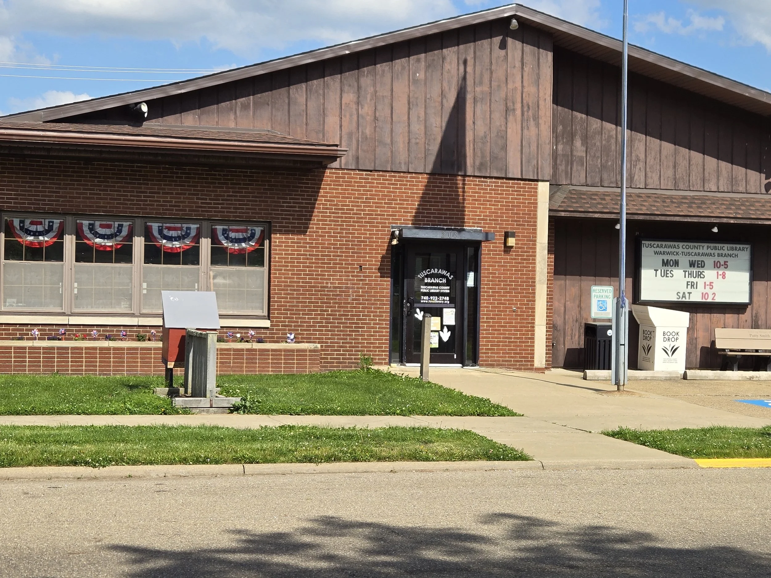 The image shows the exterior of the Tuscarawas County Public Library during daylight, with red brick and wood siding, decorated with American flag bunting, a sign of patriotic celebration. The sidewalk and a grassy area with a mailbox, bicycle rack with a bike, and a sign are visible in the foreground, while the sky is partly cloudy.