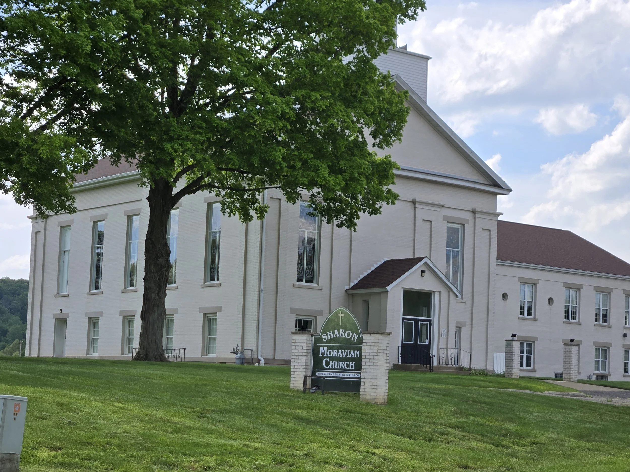 A large white church building with multiple windows, a triangular roof, and a sign in front reading 'Sharon Moravian Church' with service times. A prominent green-leafed tree with a thick trunk partially obscures the building. The sky is partly cloudy with patches of blue.