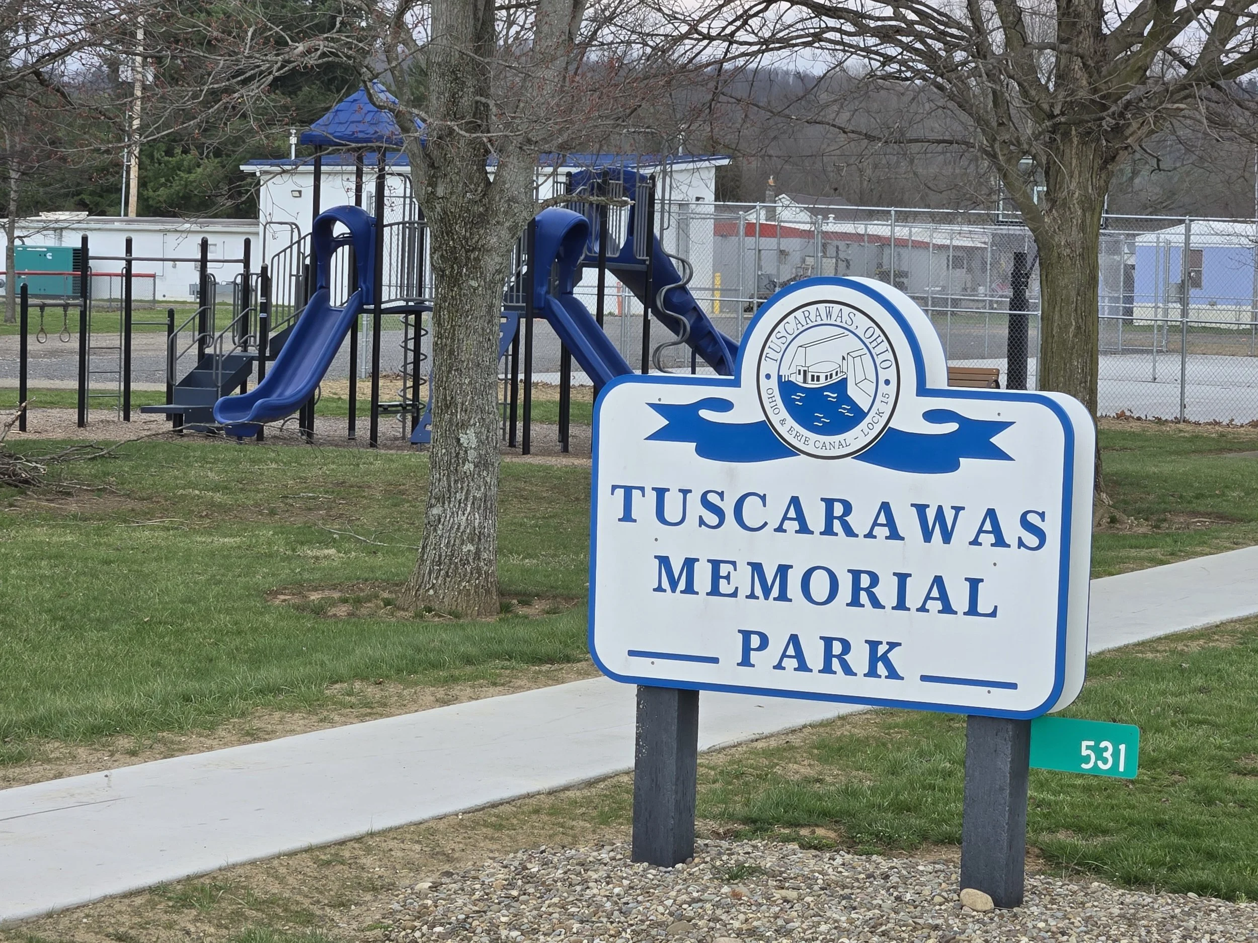 A park sign for Tuscarawas Memorial Park with a playground and trees in the background.