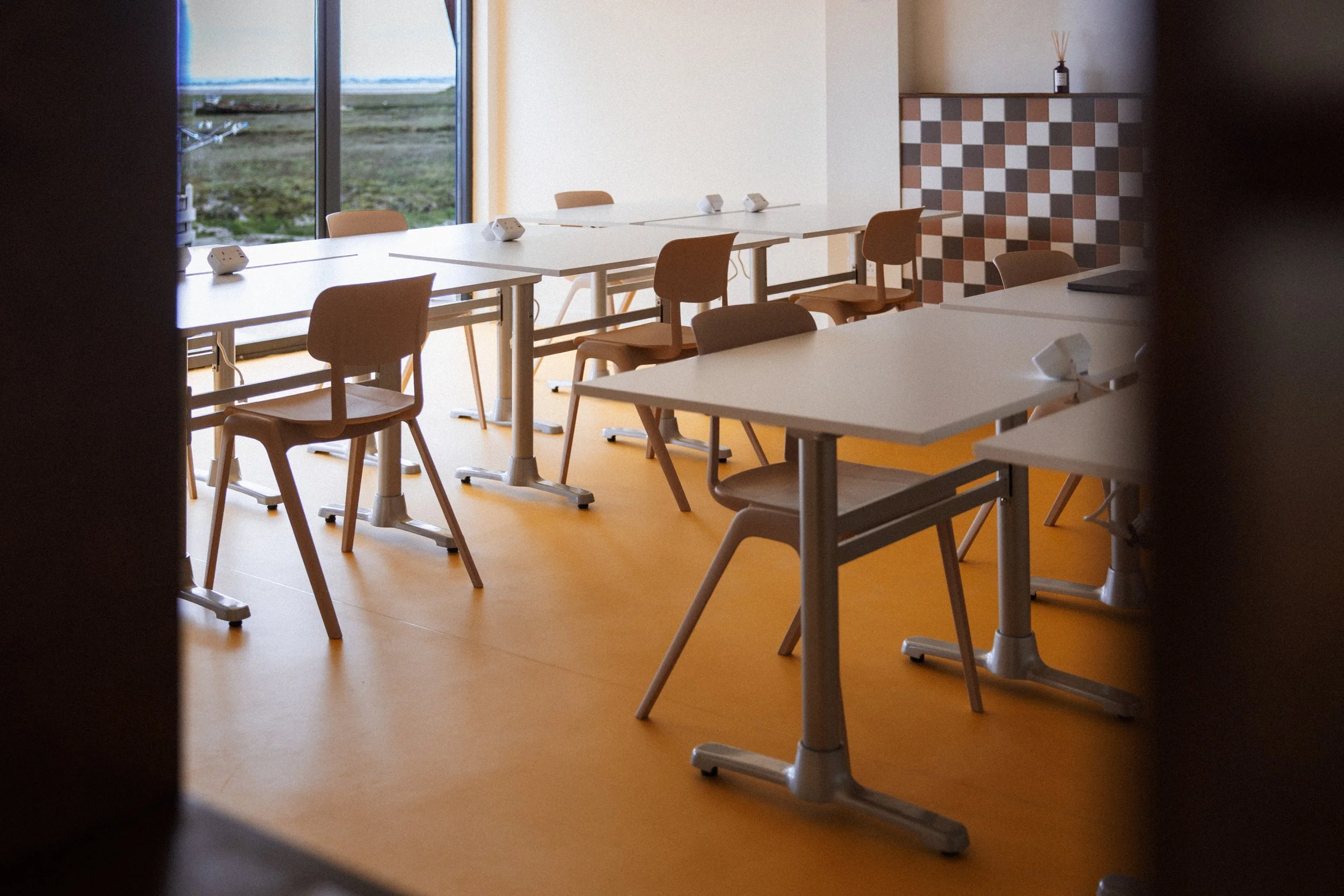A meeting room with white tables and tan chairs, large windows showing an outdoor landscape, and a decorative wall with a checkered pattern in shades of brown and gray.