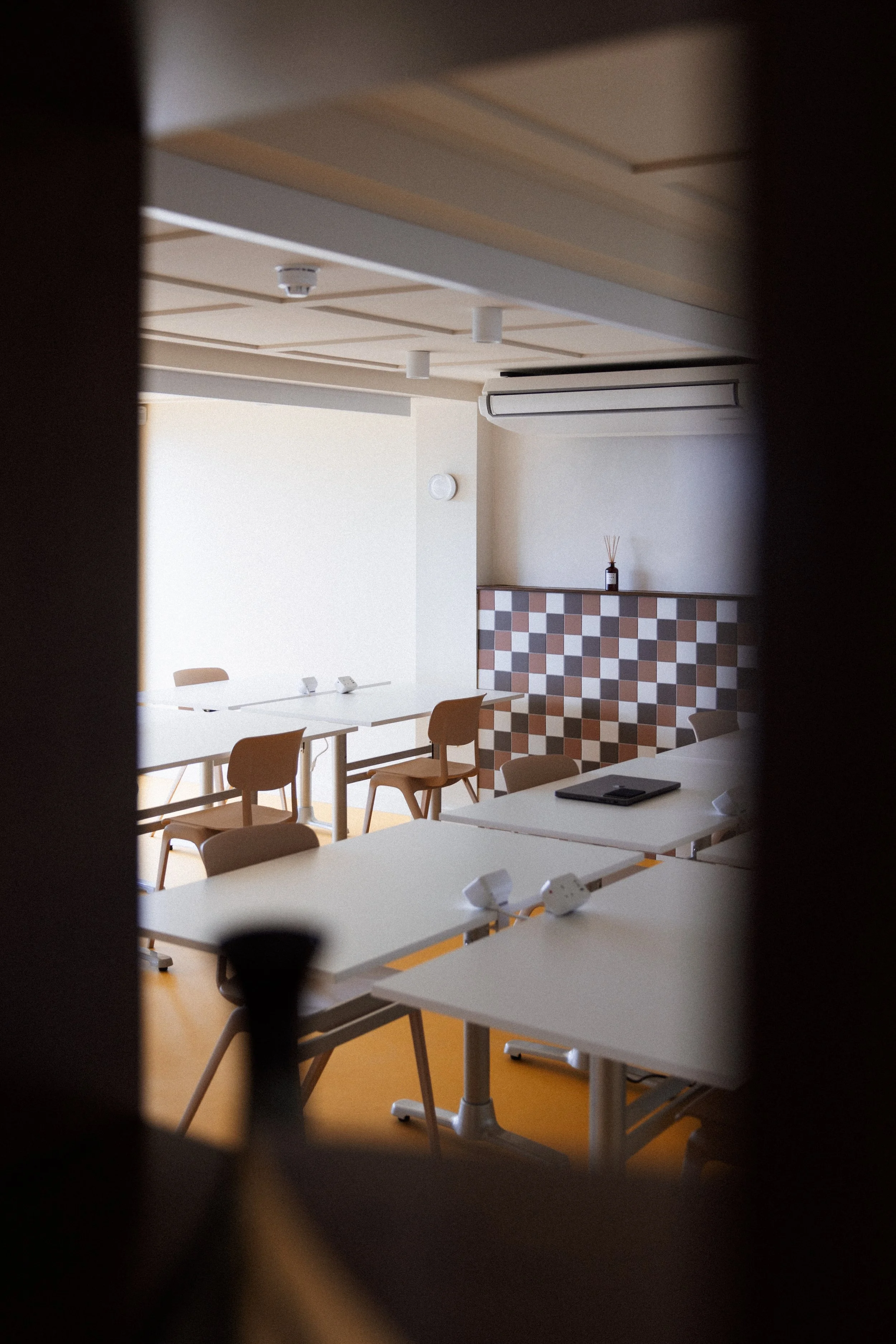 View of a modern conference or classroom room with white tables, beige chairs, a patterned divider, and an air conditioning unit, through a narrow gap.